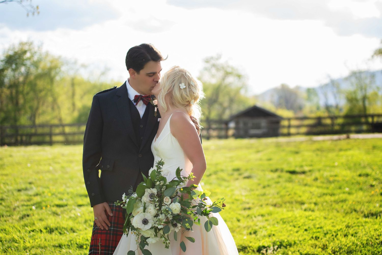 Bride and groom share a kiss in a lush green field, groom wearing a kilt, bride holding a white floral bouquet.