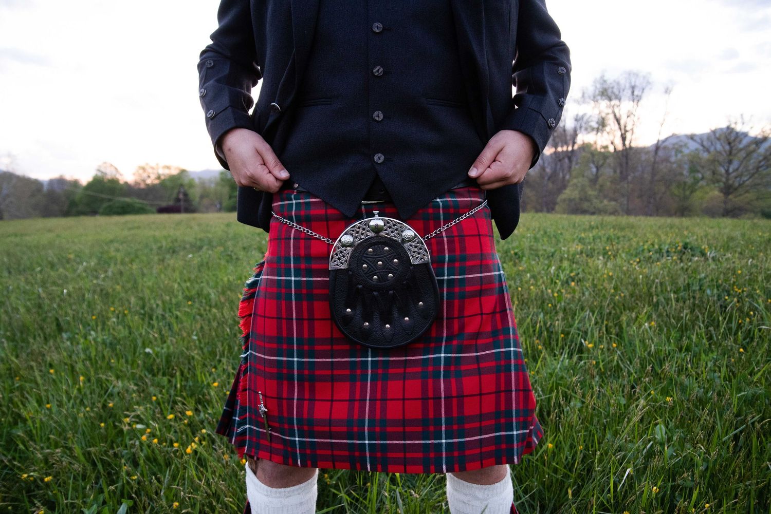 Man wearing a traditional Scottish red tartan kilt with sporran, standing in a green field at dusk.