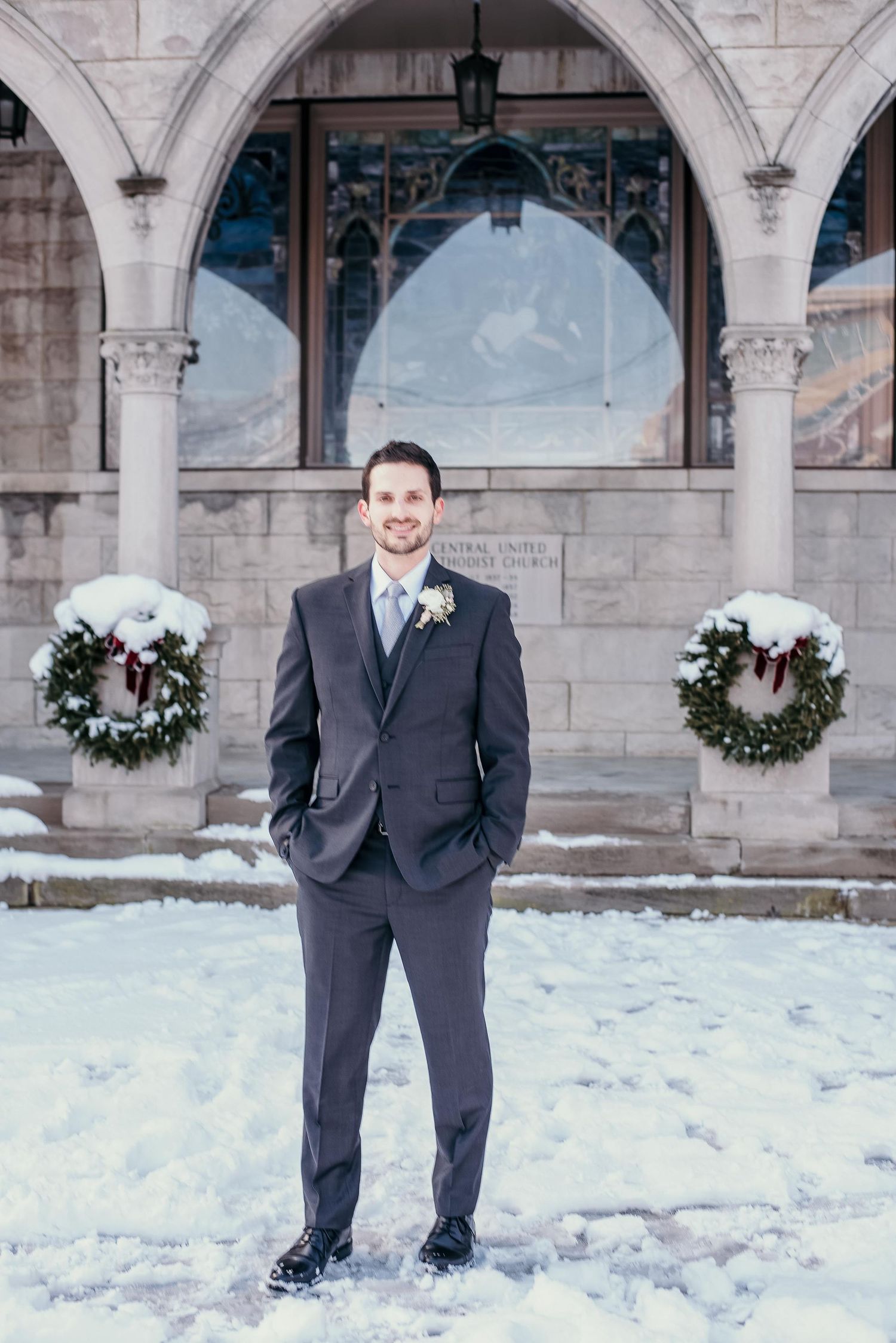 Groom in dark suit with boutonniere stands in snow outside stone church with Christmas wreaths.