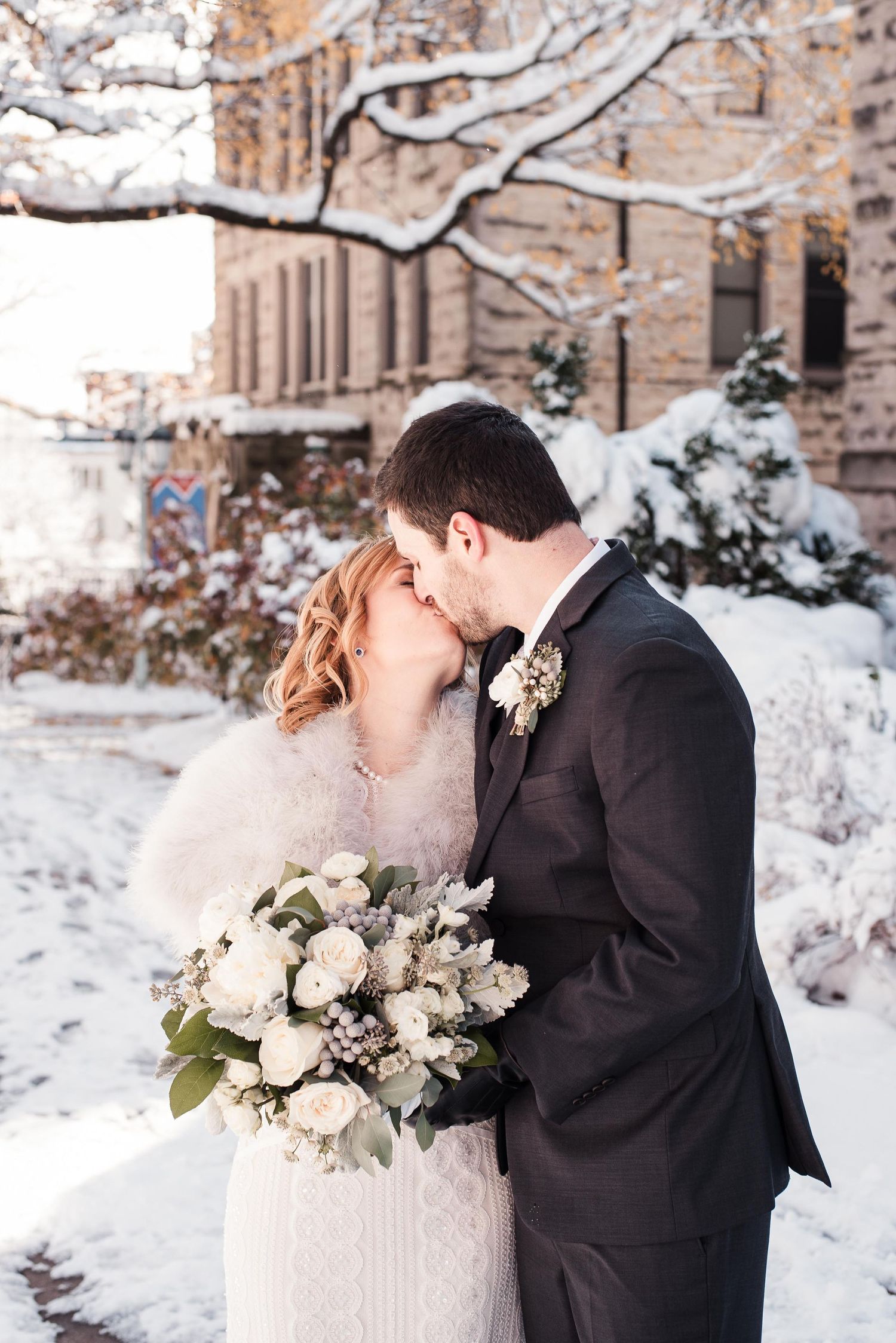 Bride and groom sharing a kiss in a snowy winter setting, bride holding a white floral bouquet with greenery.