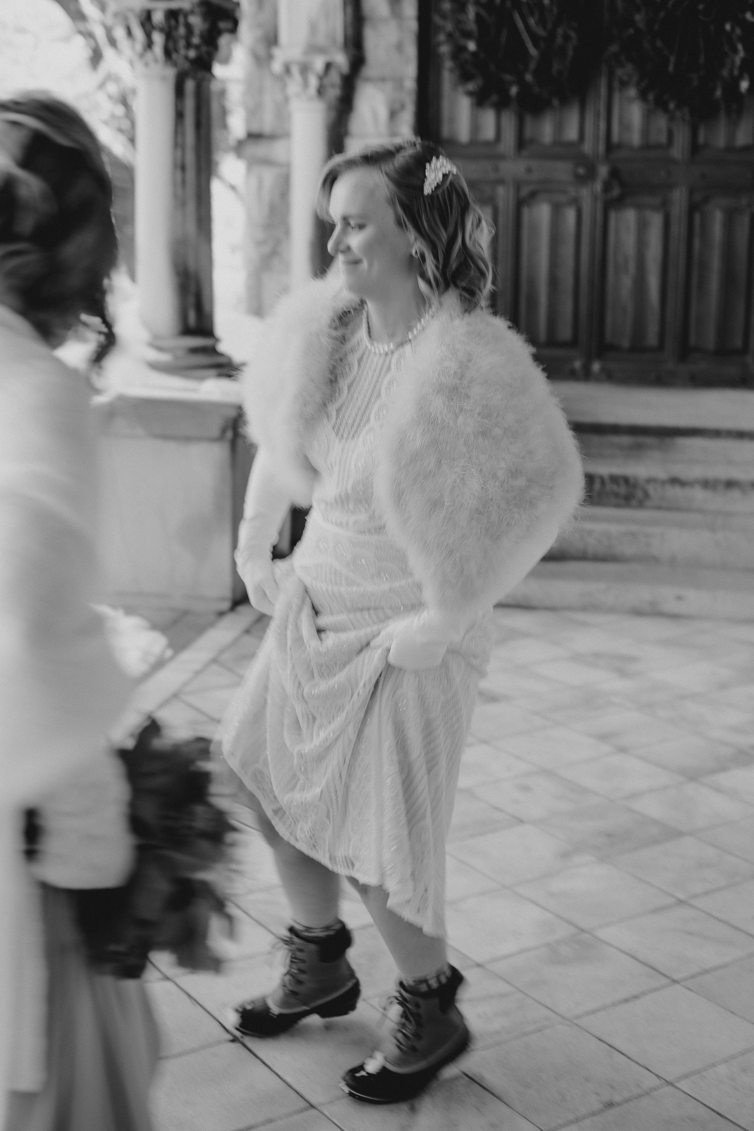 Black and white photo of a bride in a fluffy wrap, midi dress, and winter boots smiling outdoors at a classic stone venue.