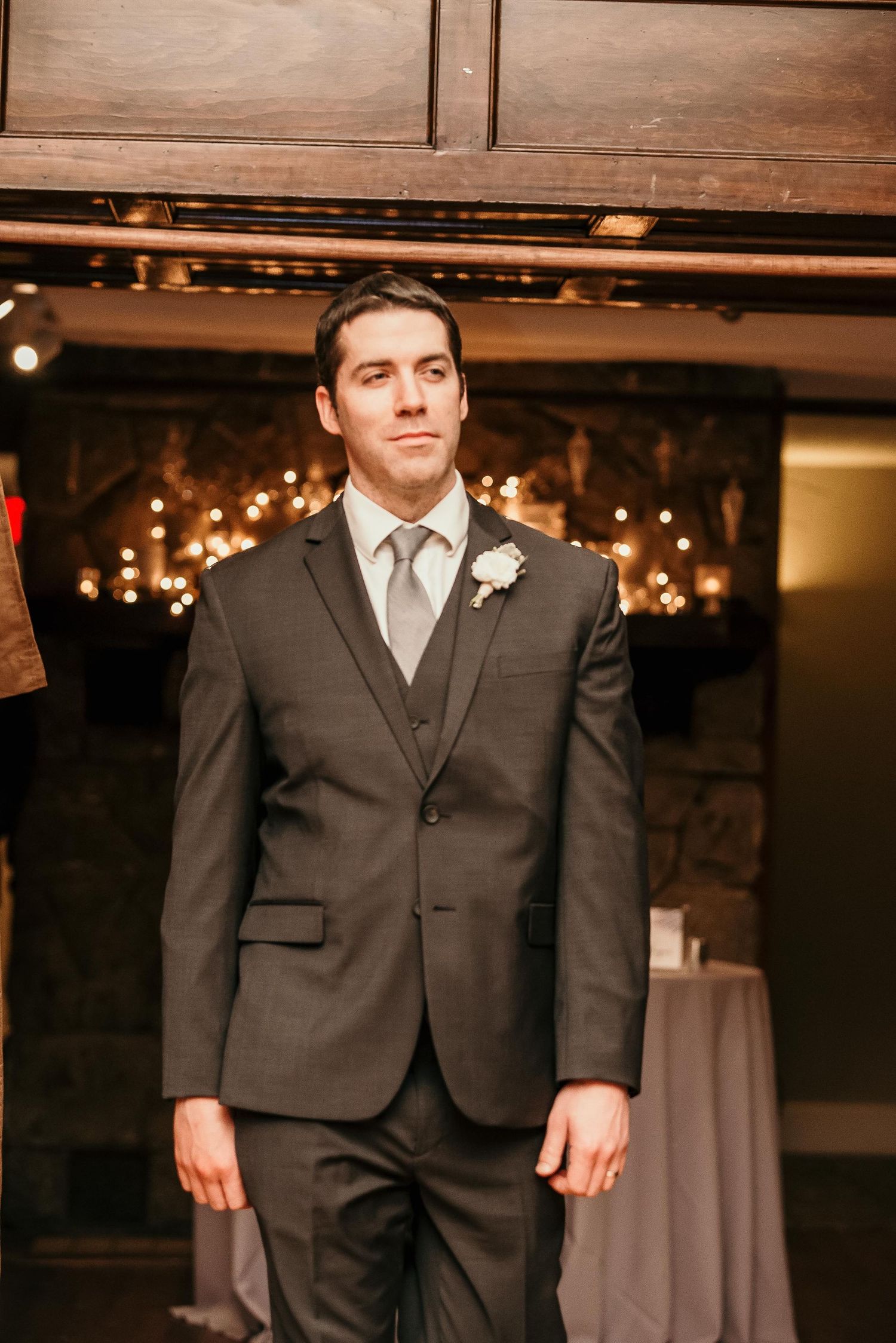 A groom in a charcoal suit with gray tie and boutonniere stands confidently at an elegant wedding venue.
