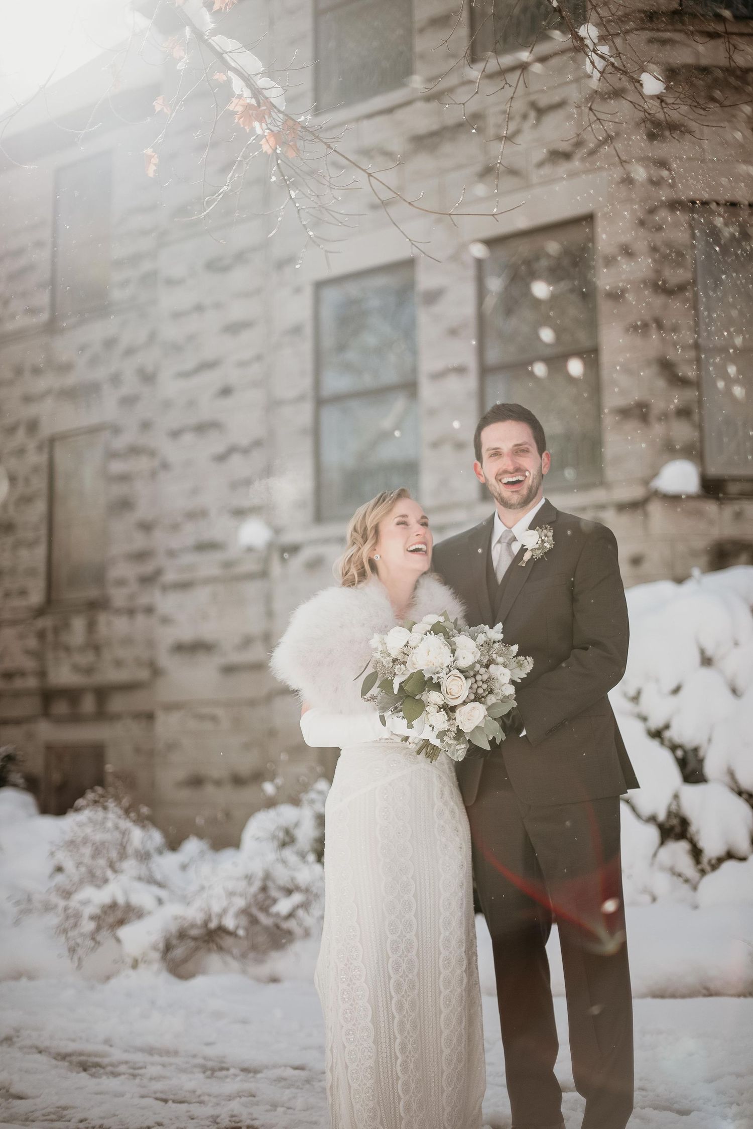 Smiling bride and groom stand together in snow outside stone building, bride holding white bouquet wearing fur wrap.