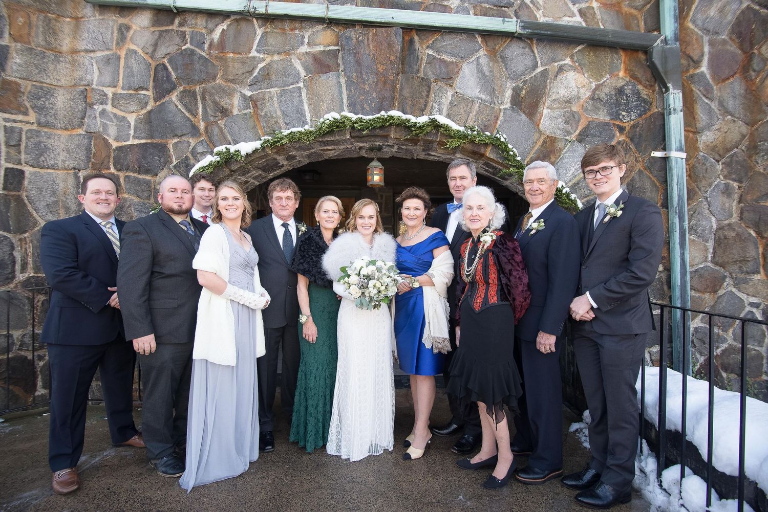 Wedding party posing in front of a stone arch entrance, dressed in formal attire including blue and white gowns.