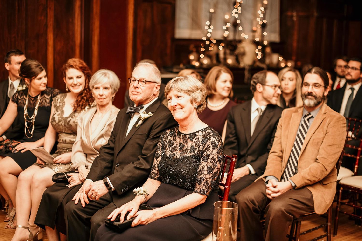 Guests smile and laugh while seated at an elegant indoor wedding ceremony with warm string lights in the background.