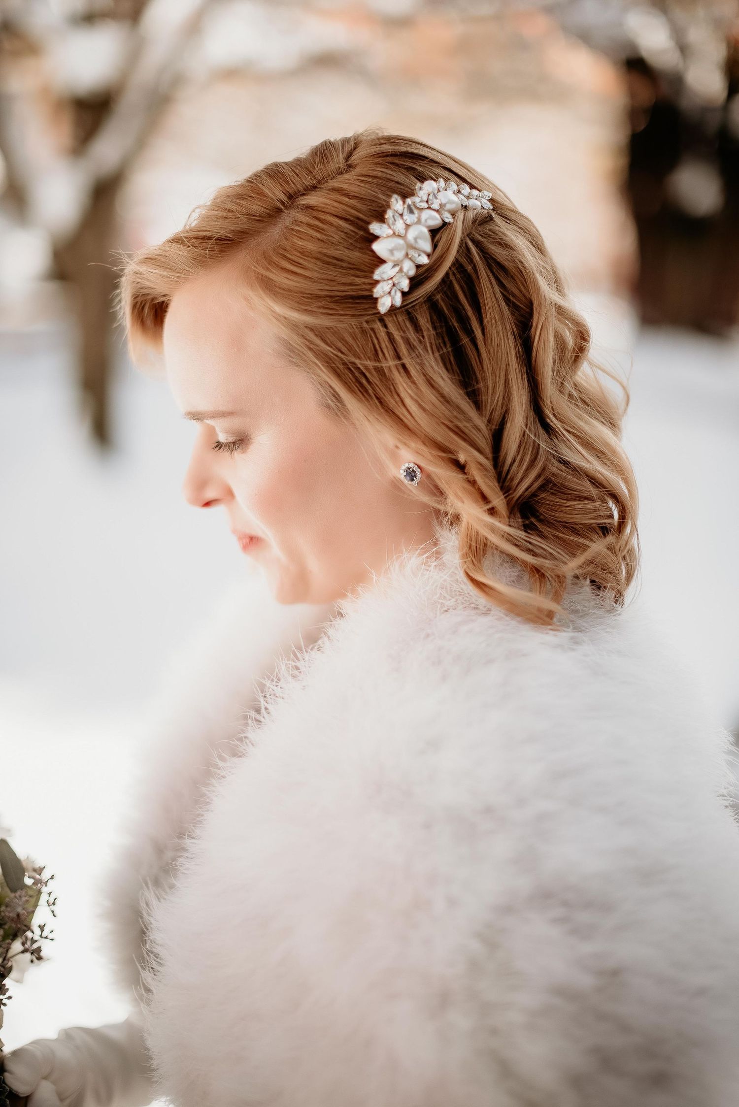 Bride in white fur wrap with jeweled hair comb, soft curls, posing outdoors in winter snow setting.