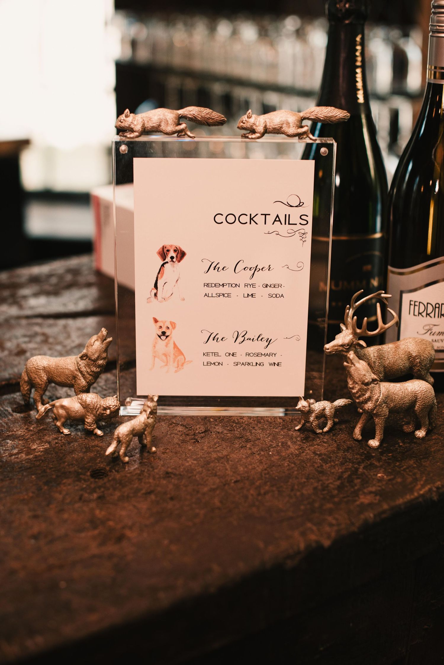 Acrylic cocktail menu with illustrated dogs, surrounded by gold animal figurines and wine bottles on a rustic bar.