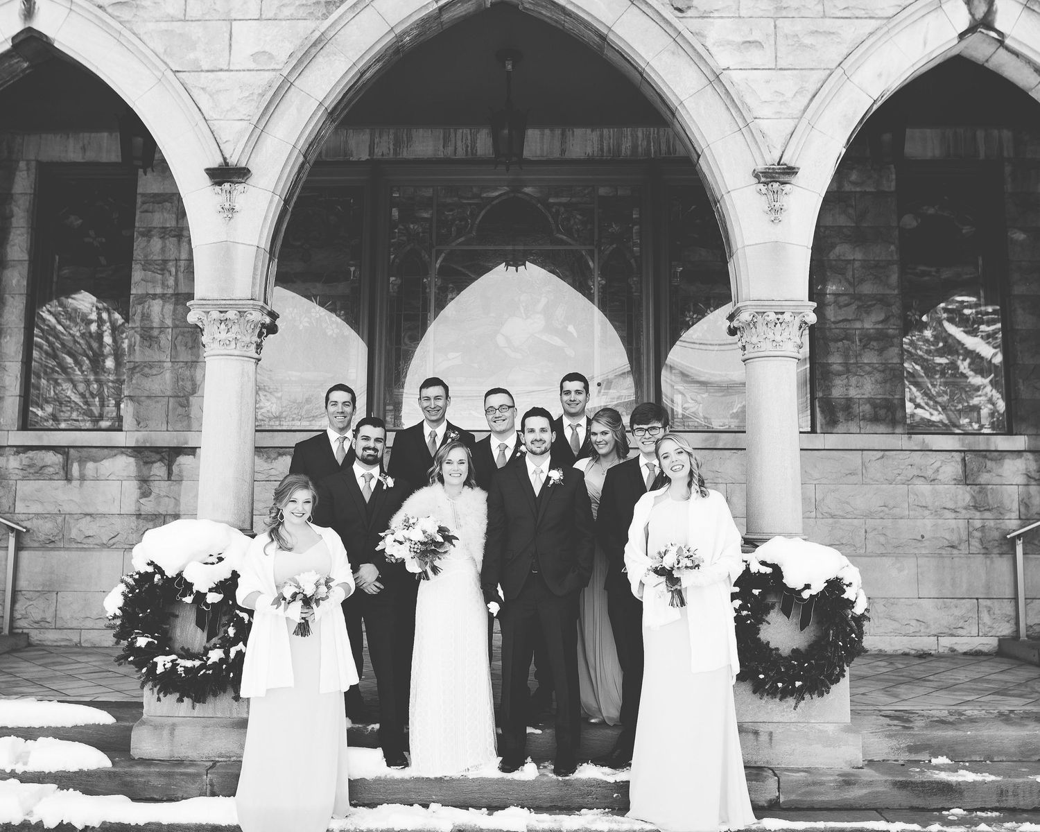 Black and white wedding party photo posed under stone arches with snow-dusted wreaths at a historic church.