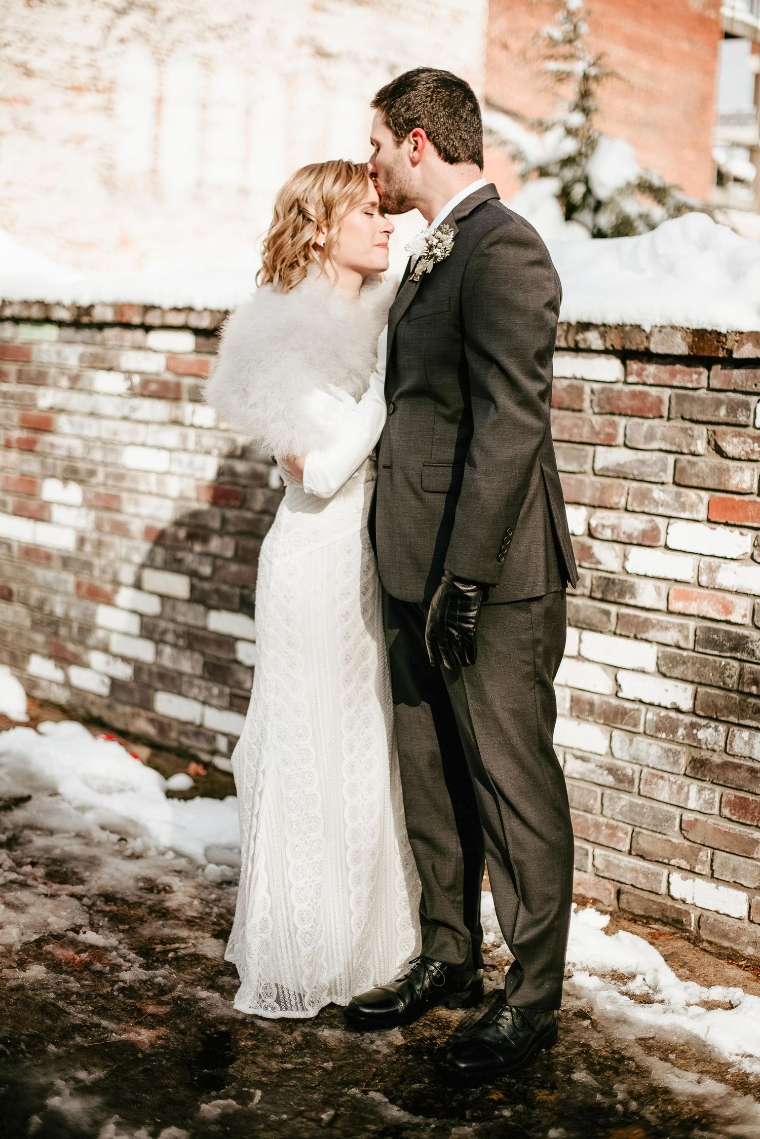 Bride and groom share a romantic winter kiss outdoors, bride in white lace gown with fur wrap, groom in dark suit.