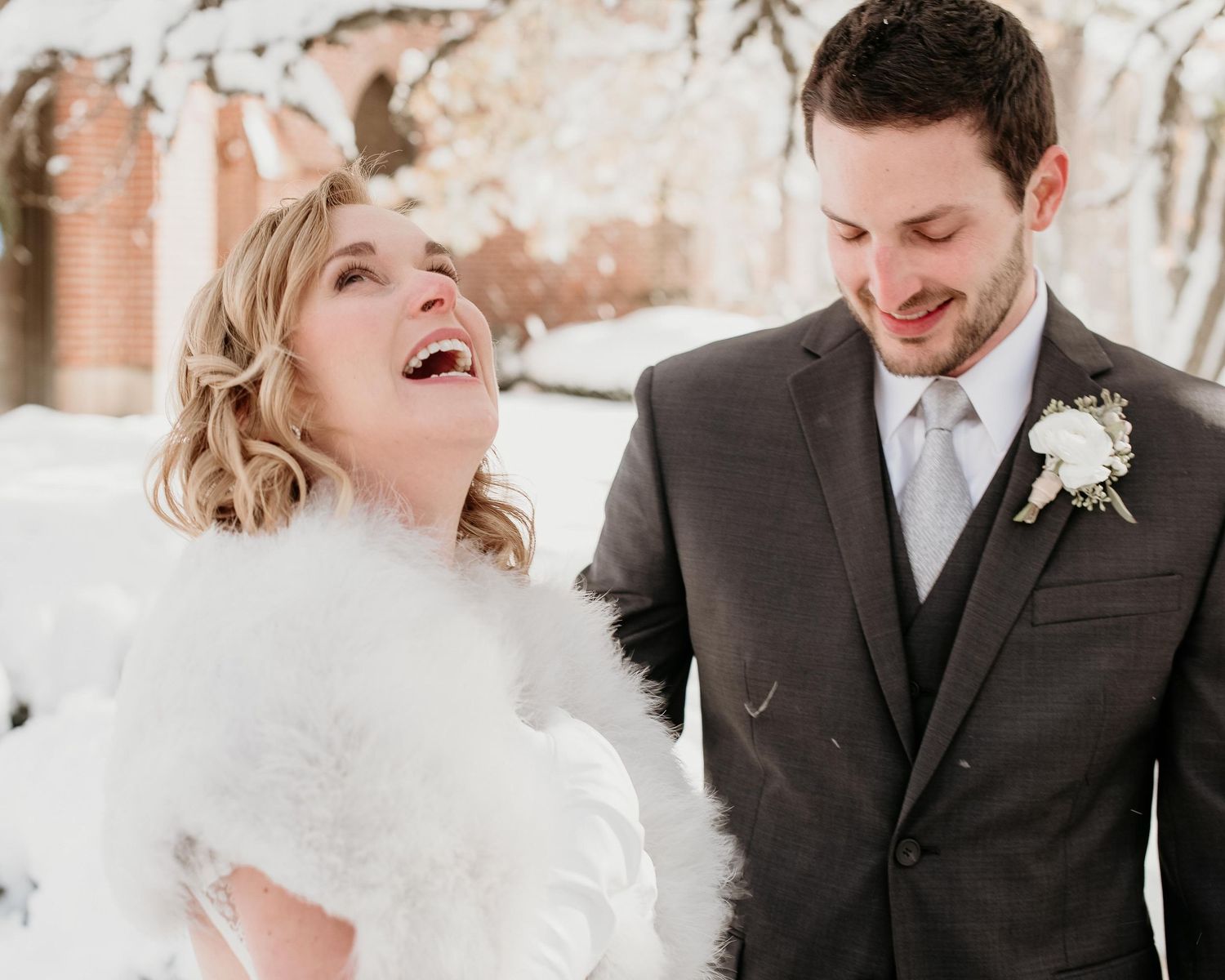A joyful bride in a white fur wrap laughs with her groom in a gray suit during a snowy winter wedding outdoors.