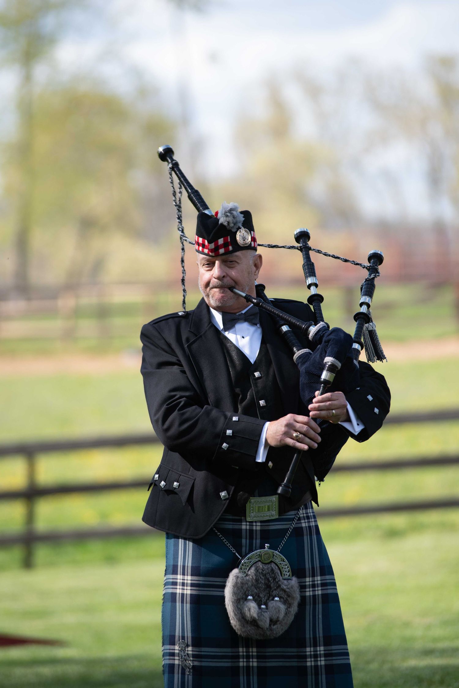 A bagpiper in traditional Scottish attire, including tartan kilt and feathered cap, plays bagpipes outdoors.