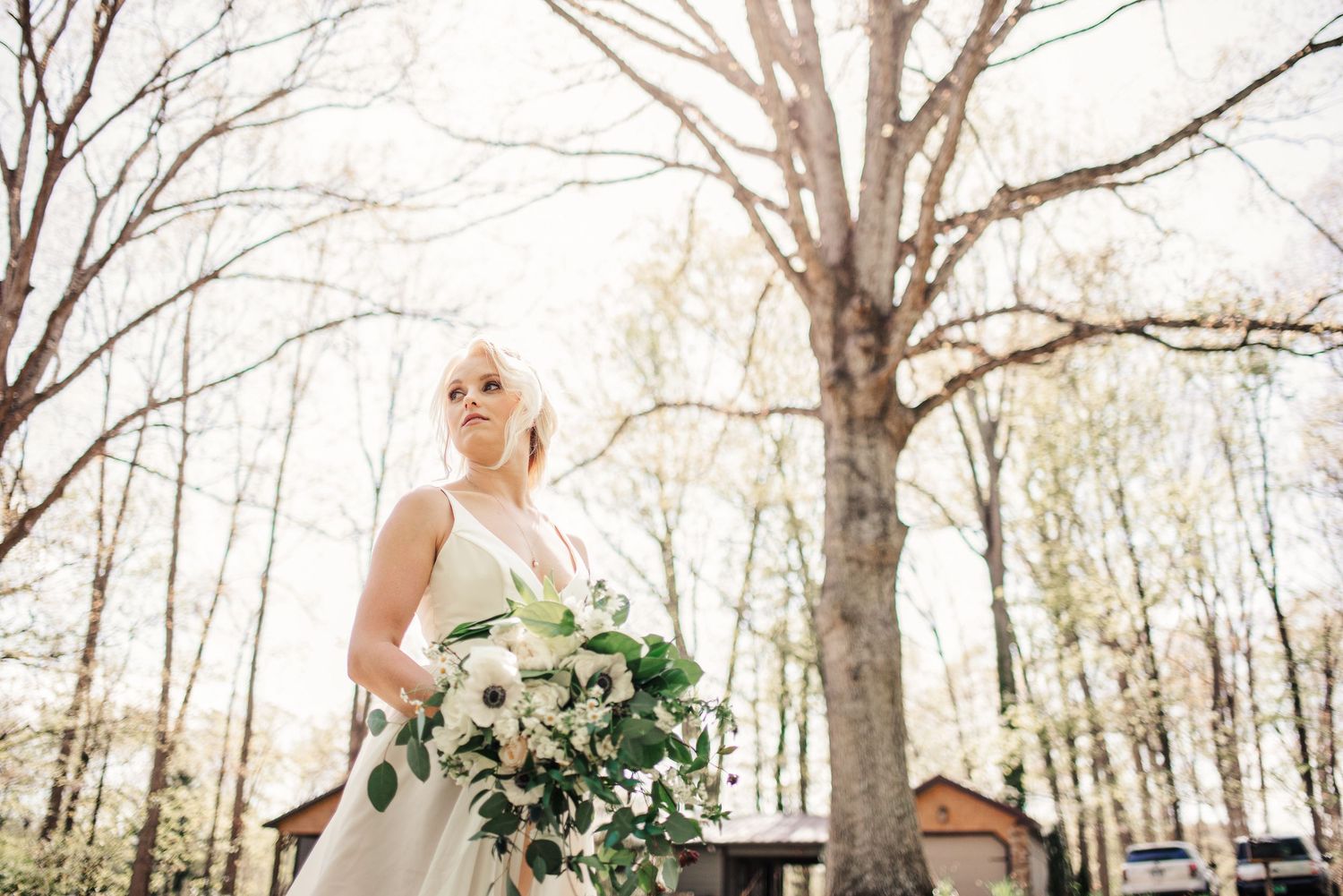 Elegant bride in white gown holds lush floral bouquet among bare trees in spring.