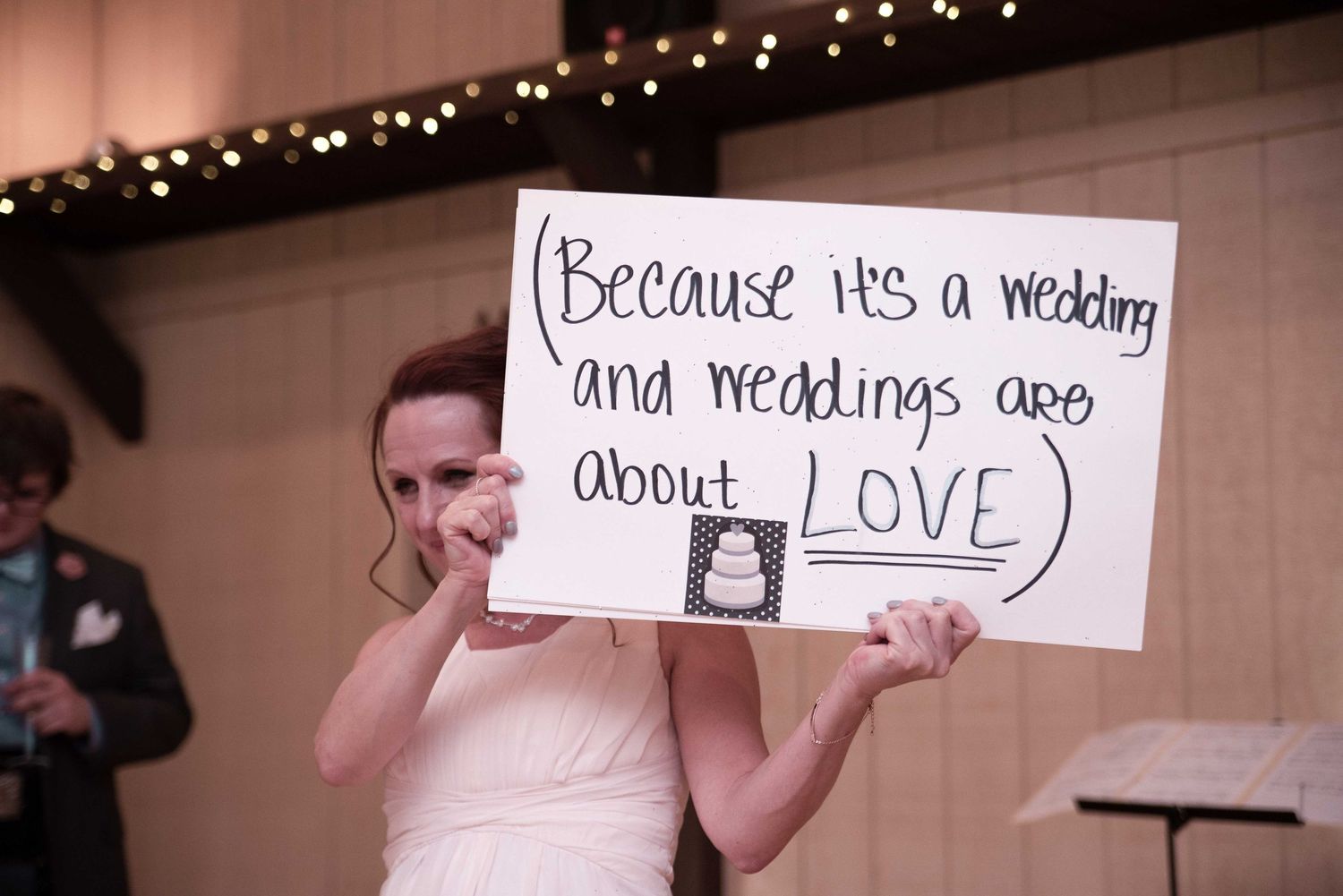 Bride holds handwritten sign reading because its a wedding and weddings are about love at reception.