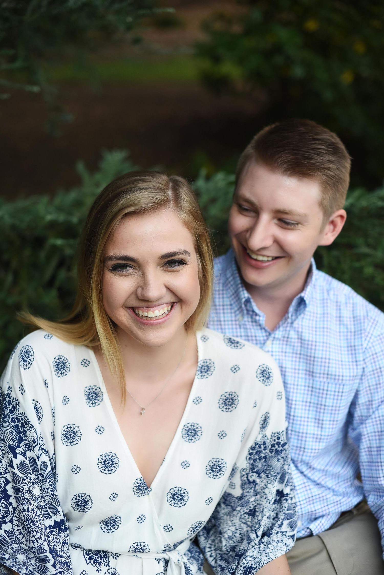 Smiling young couple posing outdoors, woman in blue floral dress with man in light blue shirt behind her.