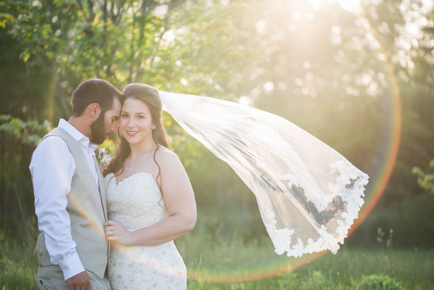 Bride smiling with flowing veil caught in wind beside groom in golden sunset outdoor wedding portrait.