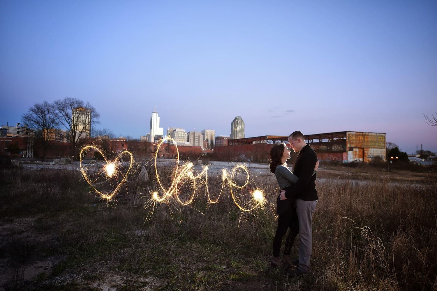 Couple kisses as sparklers spell love with city skyline at dusk.