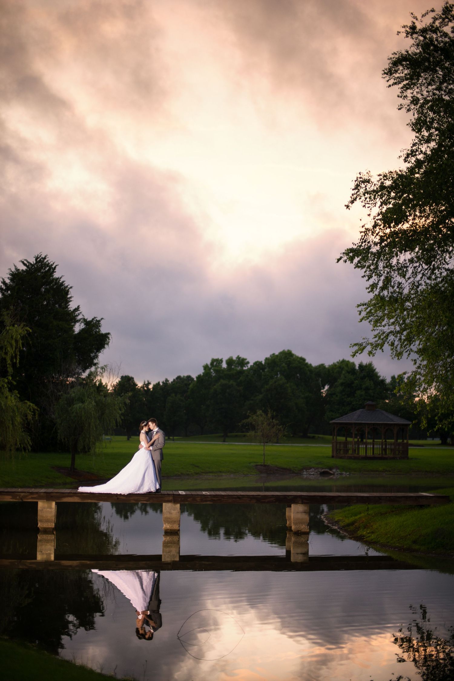 Bride and groom share a romantic kiss on a bridge reflected in a pond at sunset during their outdoor wedding.