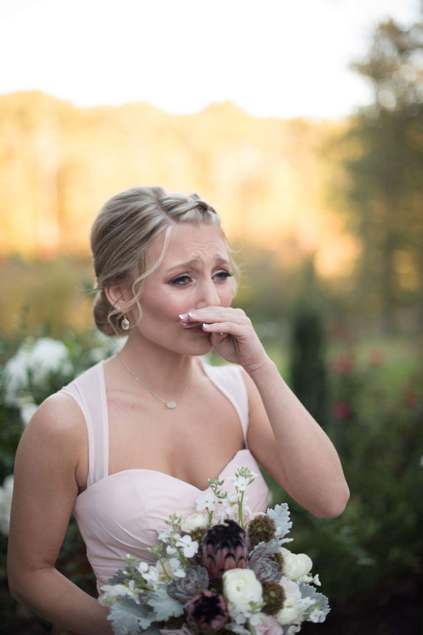 Emotional bridesmaid in blush dress wiping tears while holding a white and purple bouquet.