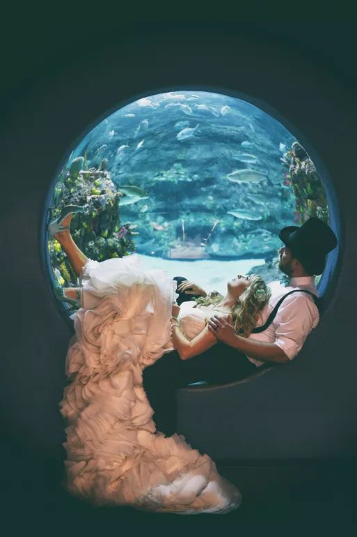 Wedding couple poses in circular aquarium window surrounded by colorful marine life underwater scene.