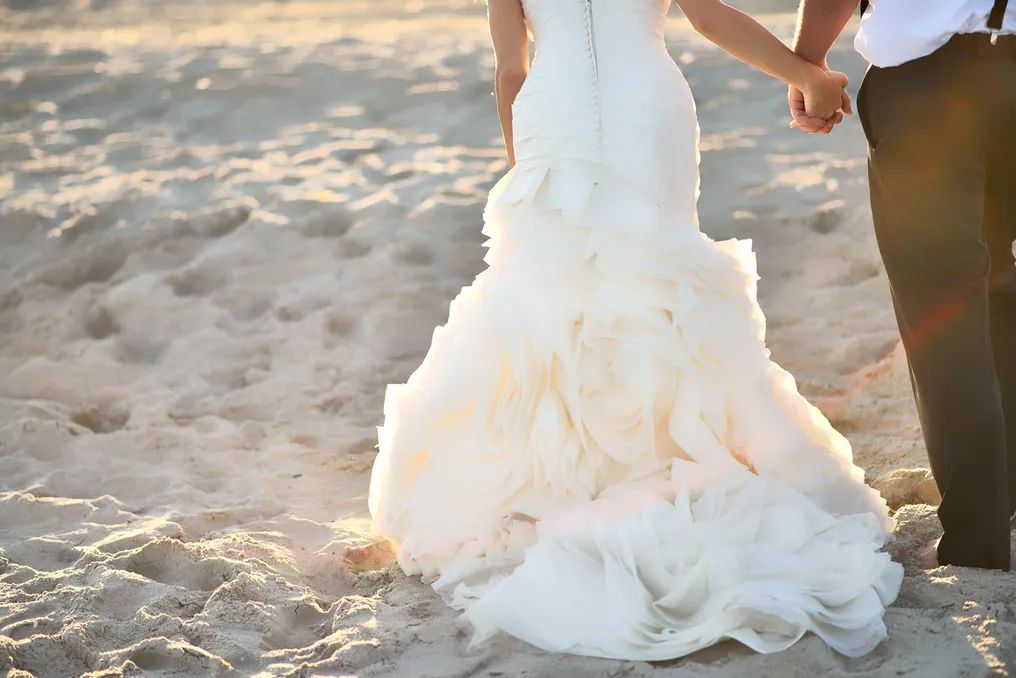 Bride in a ruffled mermaid wedding gown holding hands with groom on sandy beach.