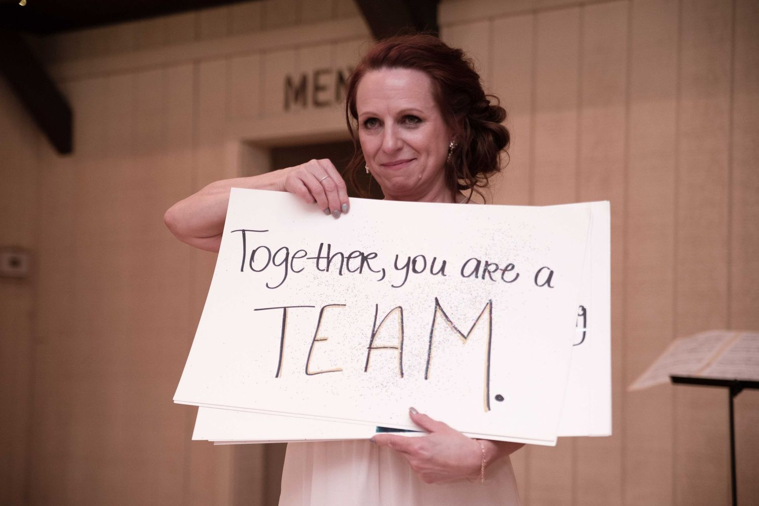 Woman holding handwritten sign reading Together you are a TEAM at indoor wedding venue.