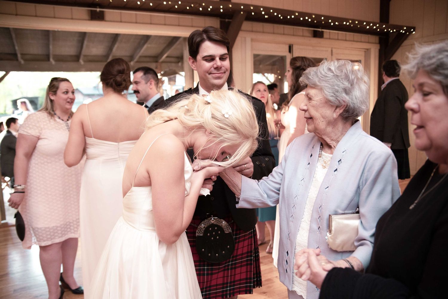 Elderly woman kisses bride's hand at wedding reception while groom in kilt smiles behind her.