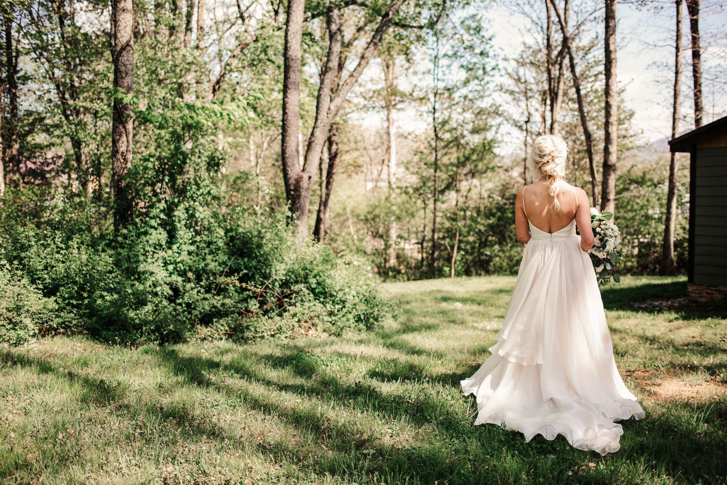 Bride in elegant backless white wedding gown standing in lush green wooded garden landscape.