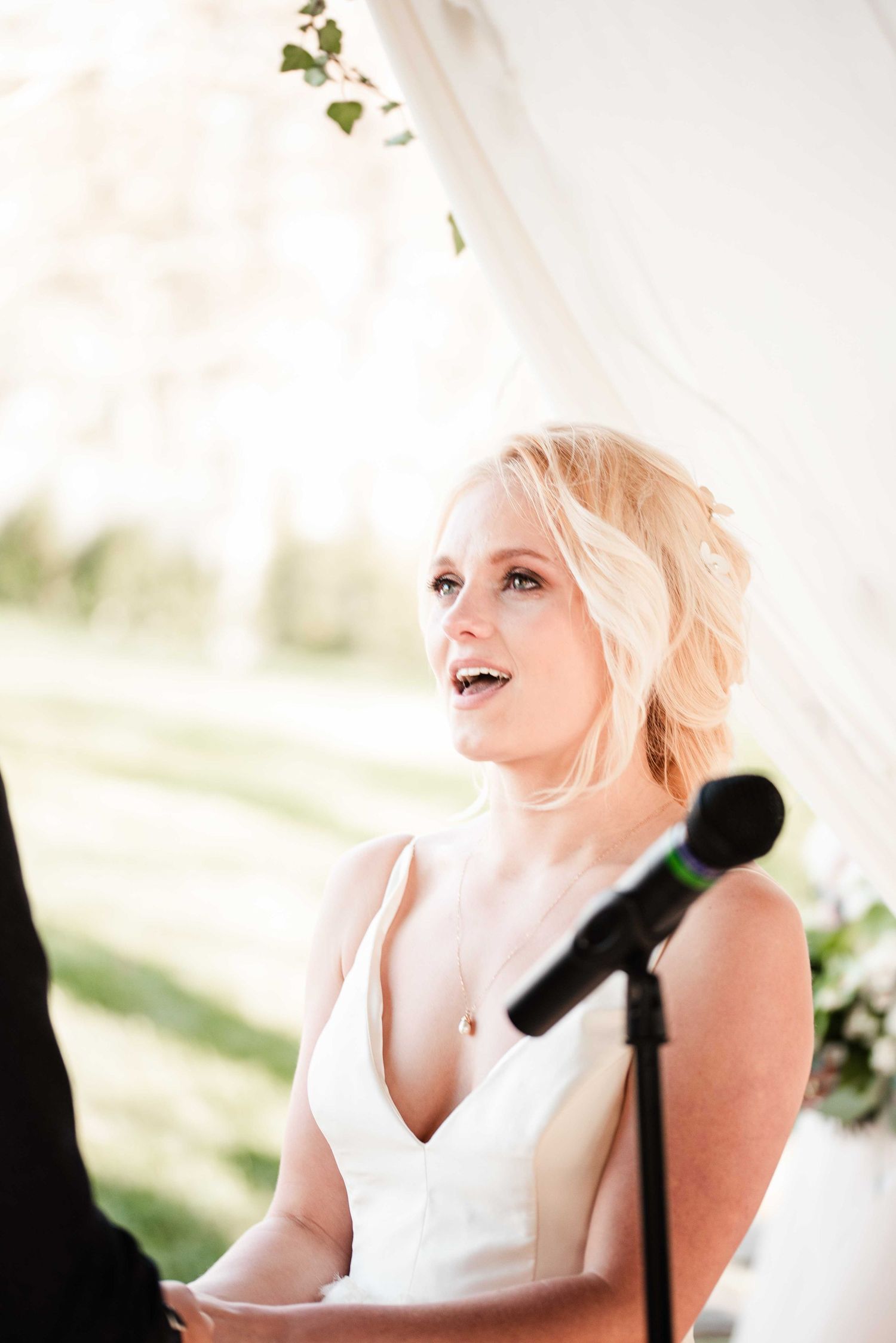 Emotional blonde bride in white V-neck gown reciting wedding vows at outdoor ceremony.
