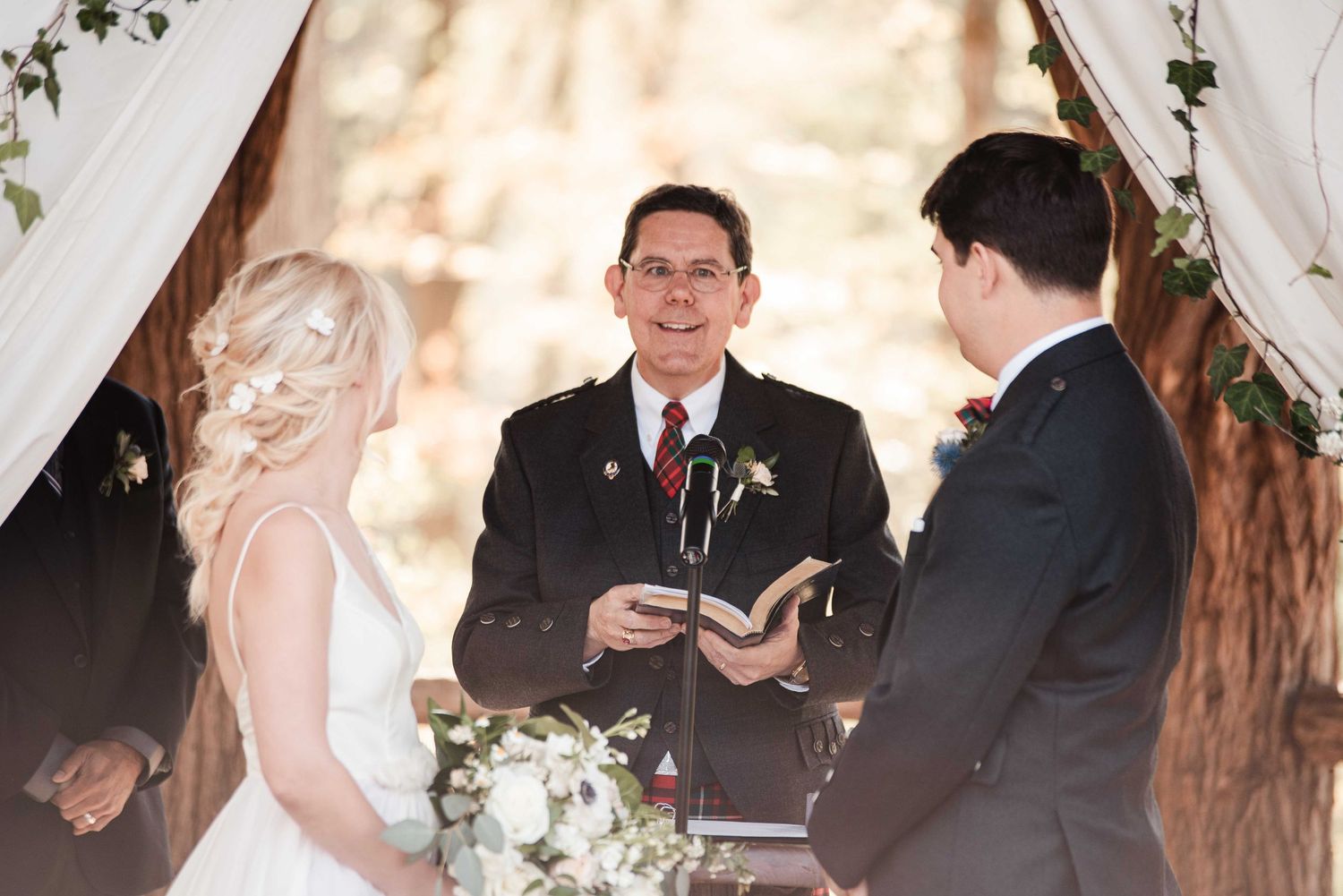 Officiant reads from a book during an outdoor wedding ceremony with bride and groom standing beneath a floral arch.