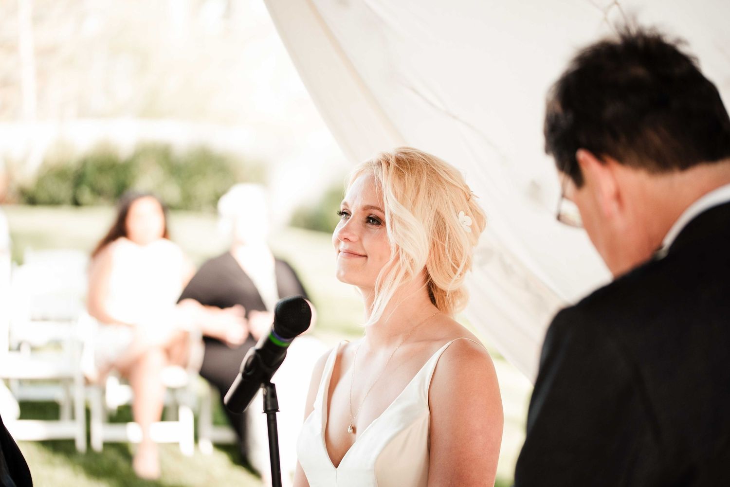 Smiling blonde bride in white gown at outdoor wedding ceremony under tent, exchanging vows at microphone.