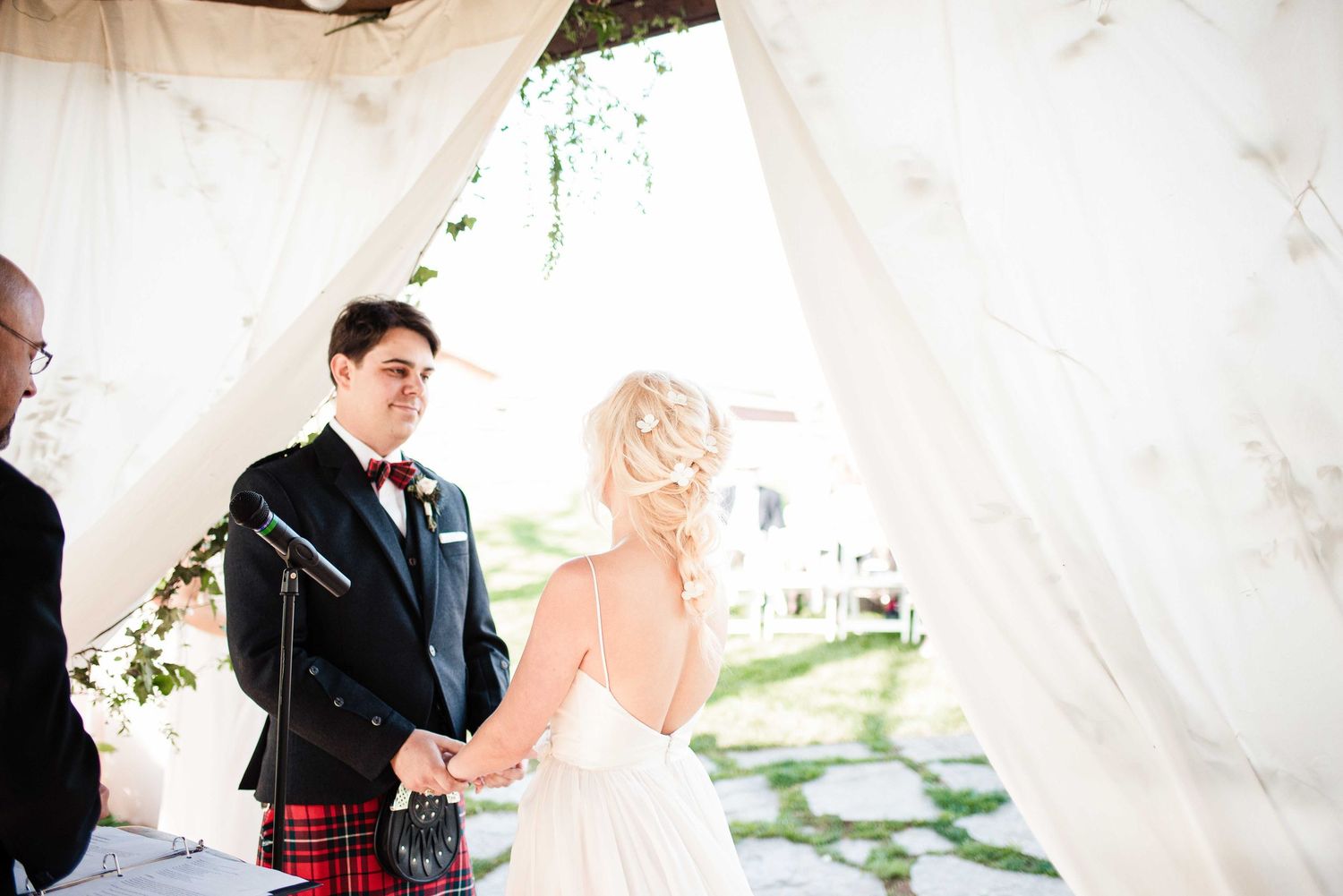 Bride and groom holding hands at outdoor wedding ceremony, groom wearing tartan kilt and red bow tie under white draped canopy.