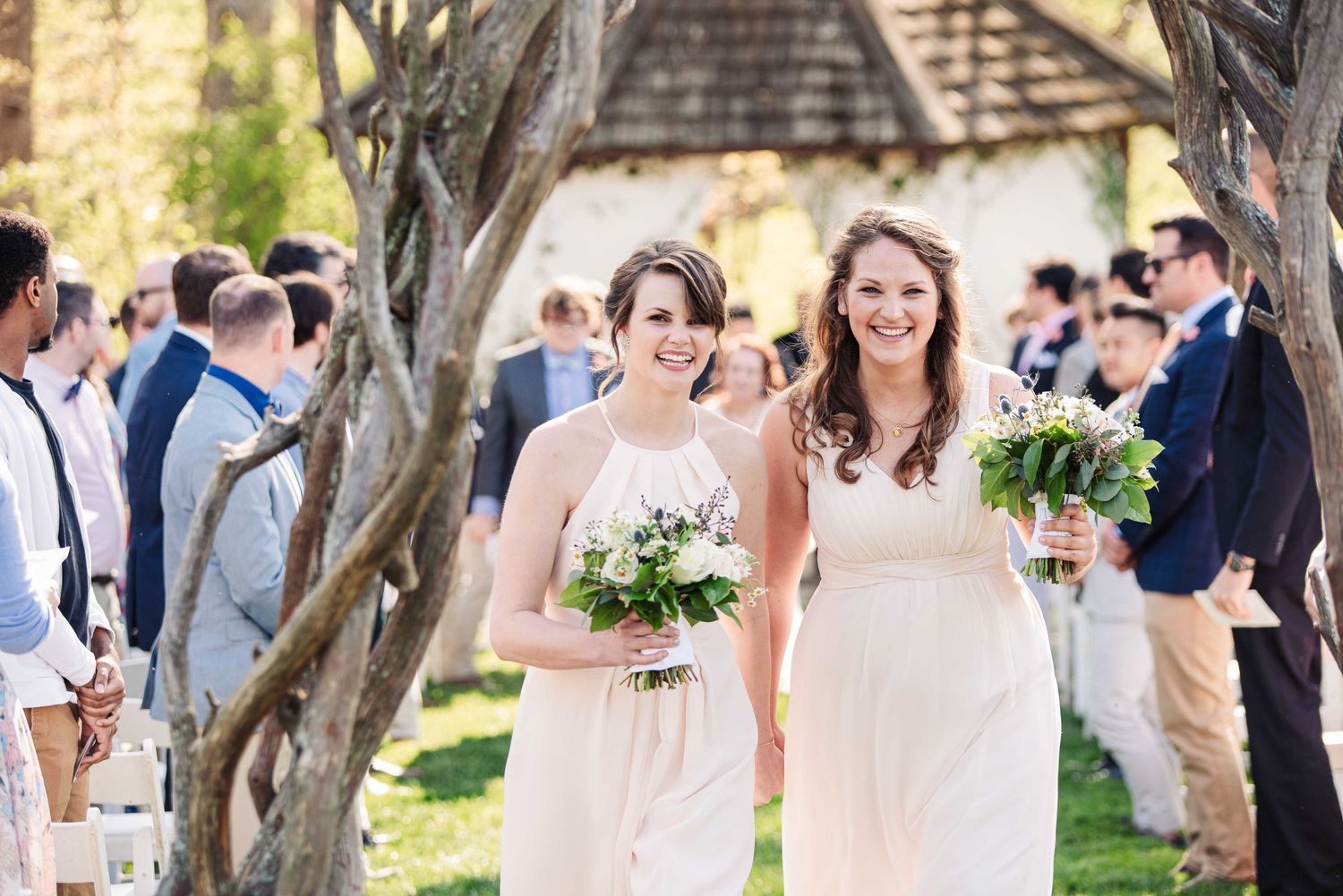 Two bridesmaids in white dresses carrying bouquets walk down outdoor wedding aisle surrounded by guests.