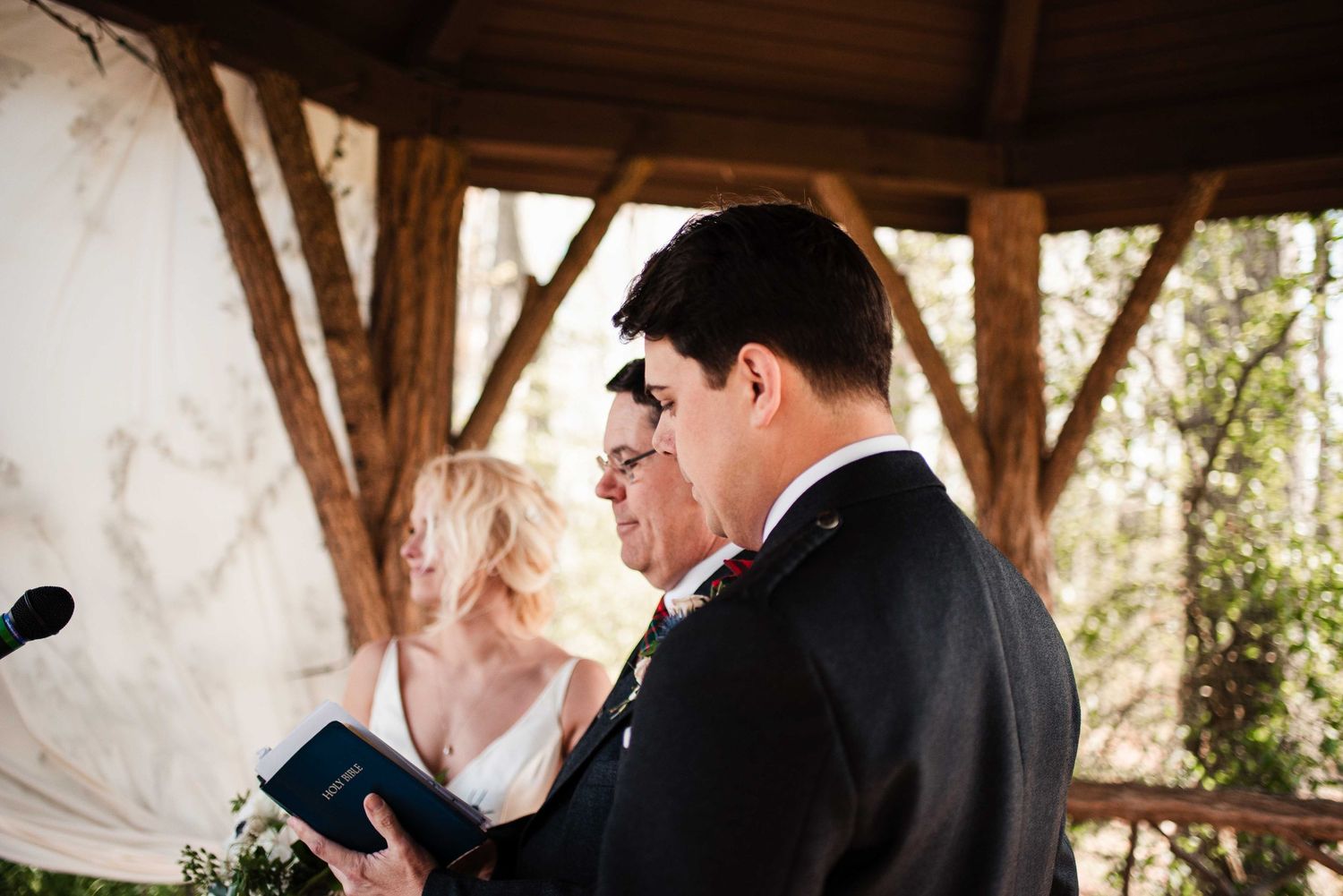 Groom reading vows at outdoor wedding ceremony under rustic wooden gazebo with bride standing behind him.