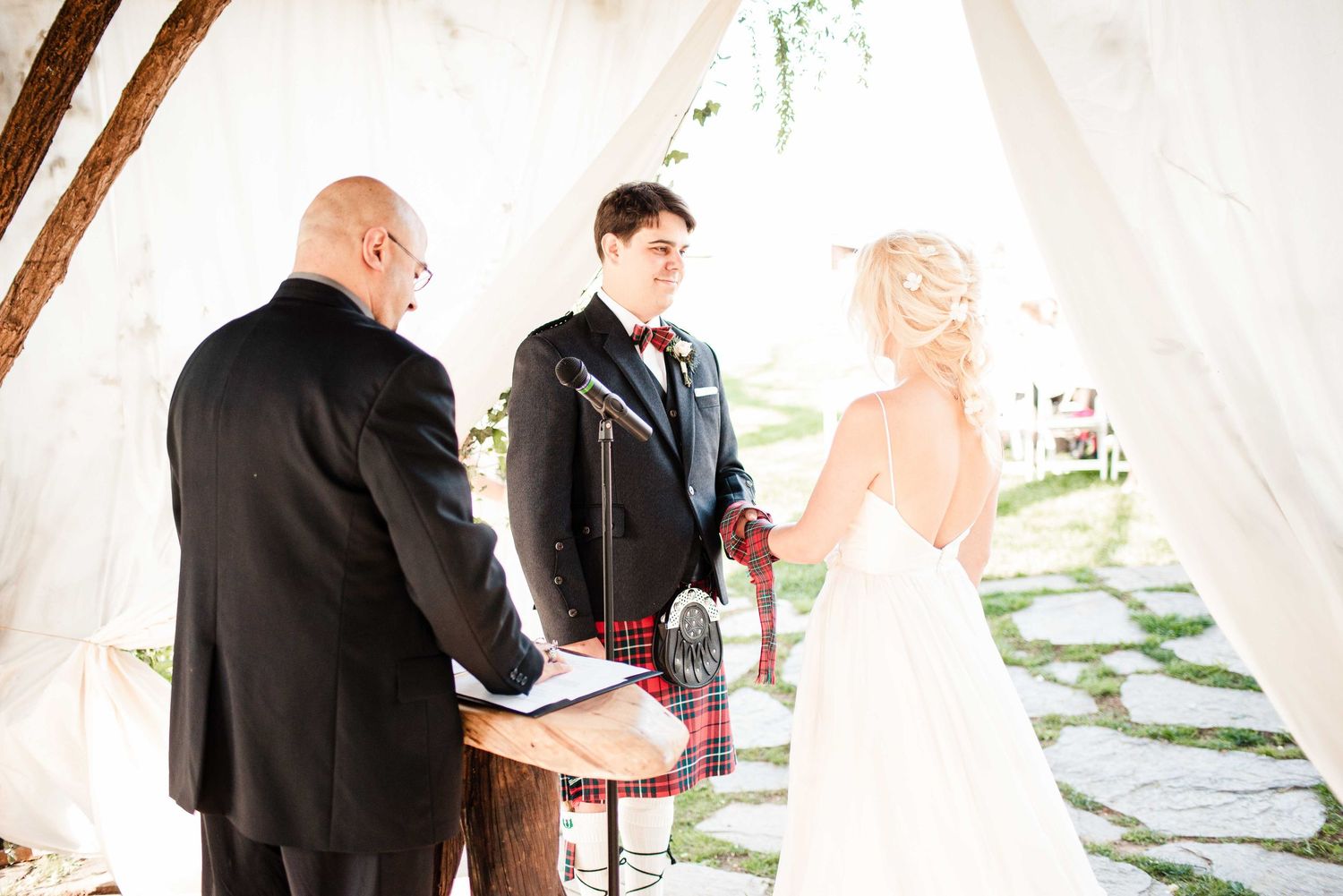 Outdoor wedding ceremony with groom in kilt, bride in white gown, and officiant under draped white canopy.