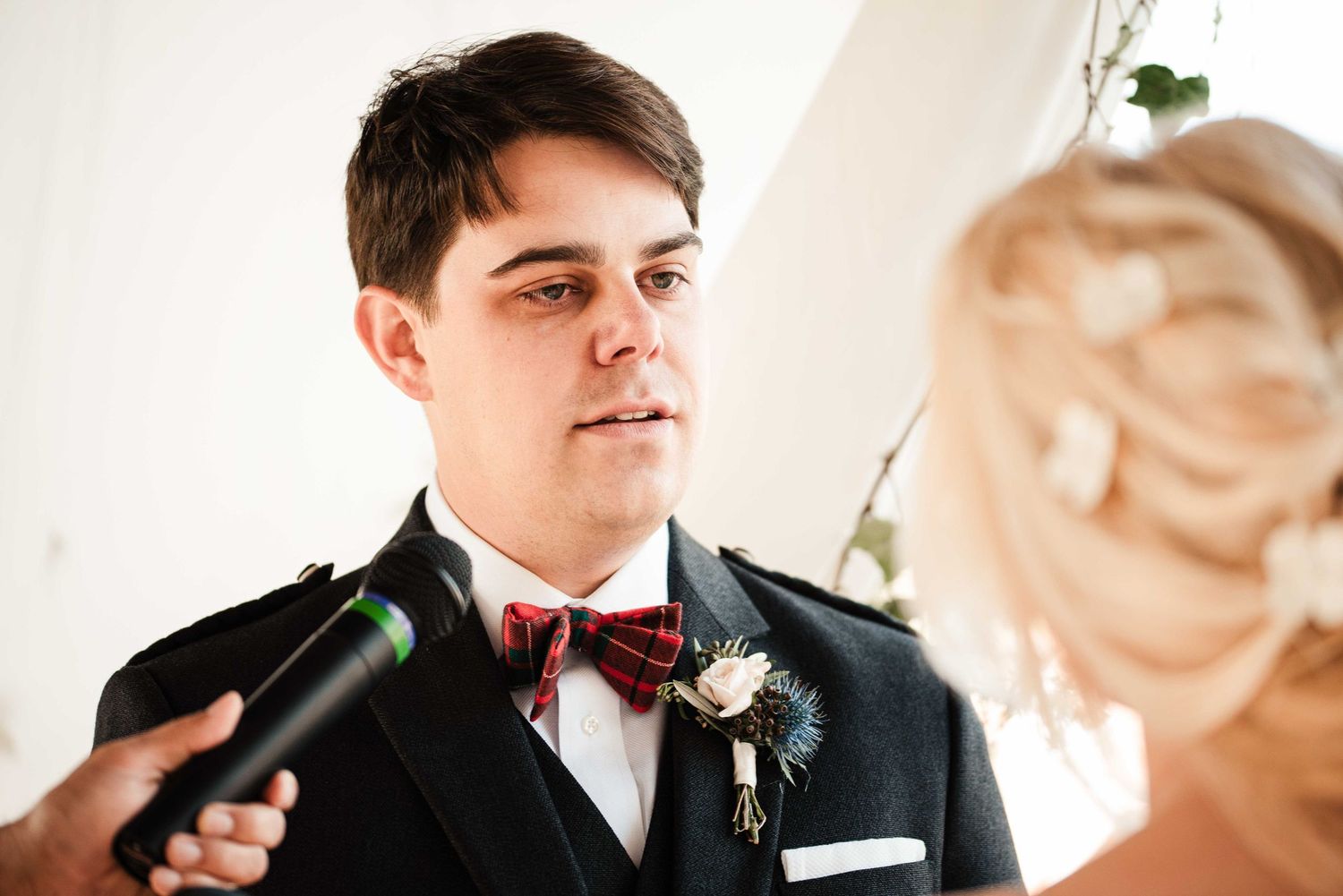 Groom in black suit with red bow tie and floral boutonniere listens attentively during wedding ceremony.