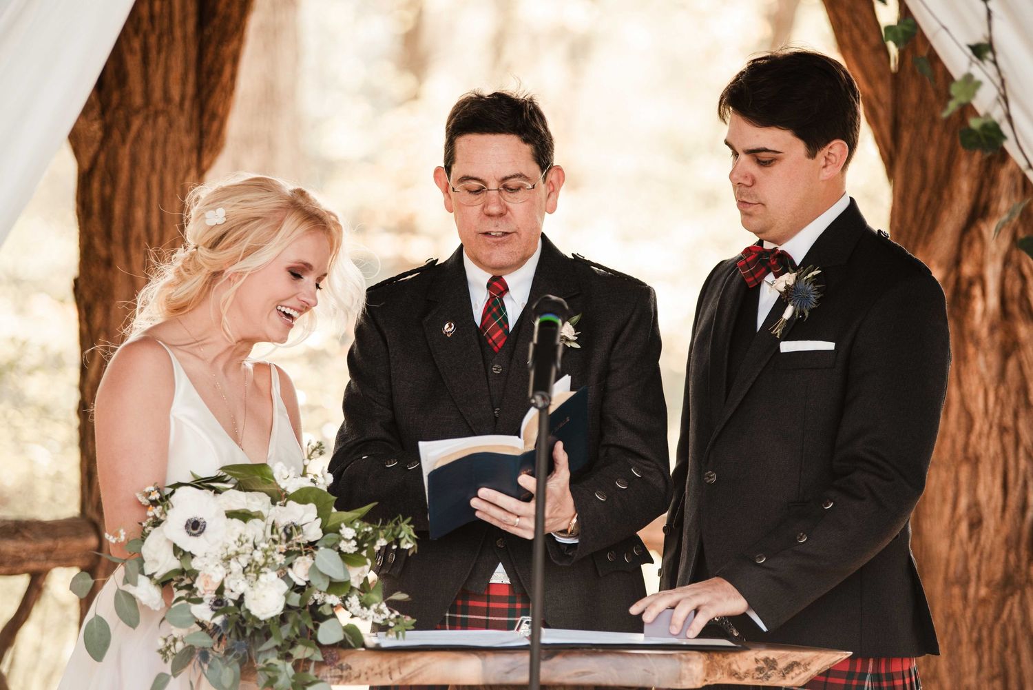 Bride and groom exchange vows with officiant in tartan kilt at an outdoor wedding ceremony.