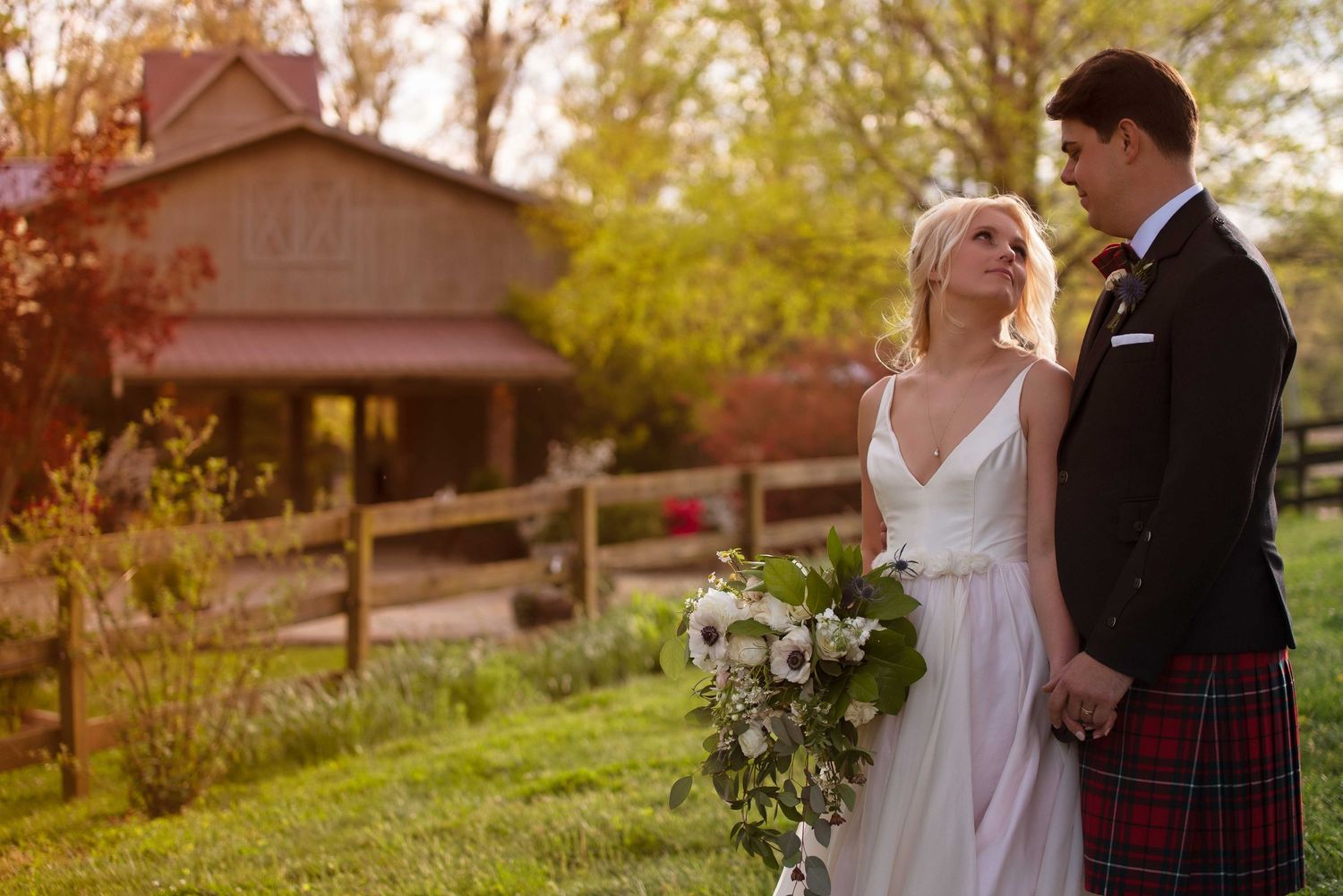 Bride and groom holding hands in a lush green field, with a rustic red barn in the background during golden hour.