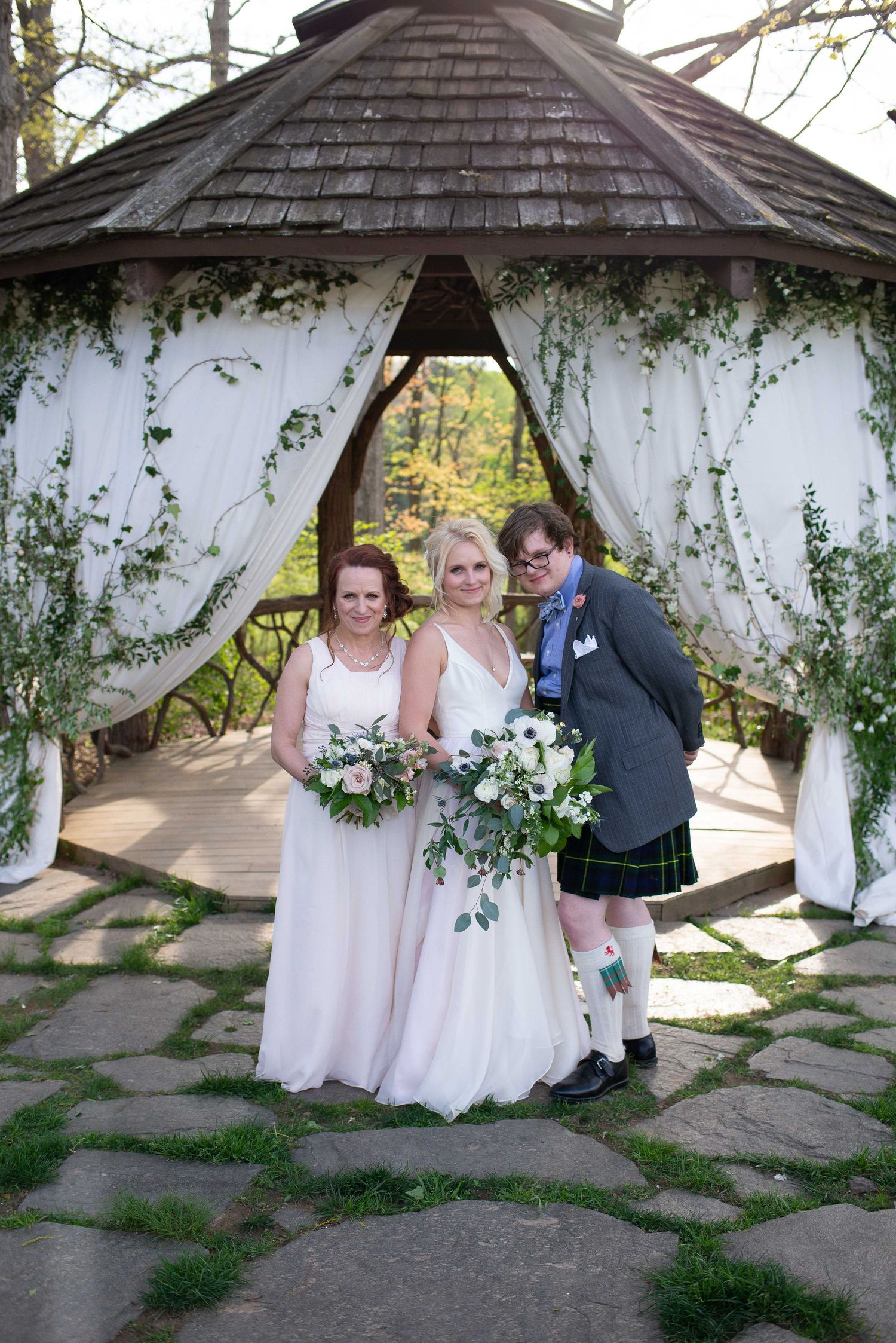 Two brides in white gowns and a guest in a kilt pose under a floral gazebo at an outdoor wedding ceremony.