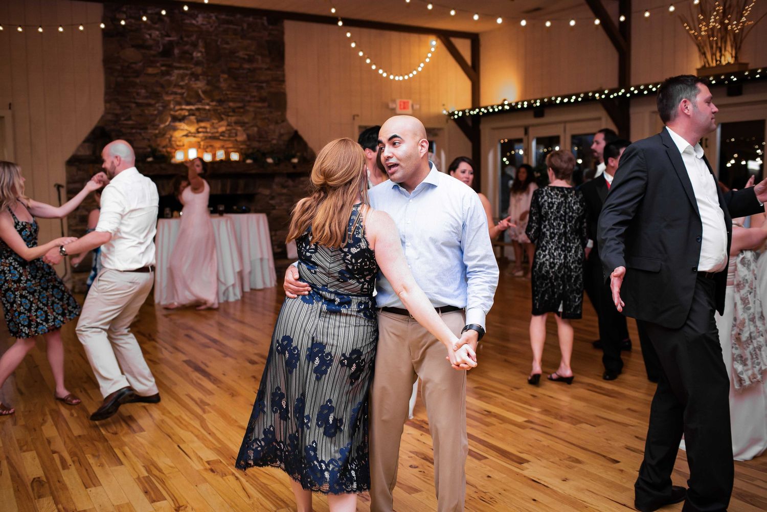 Guests dancing joyfully at a rustic wedding reception on a hardwood floor with string lights overhead.