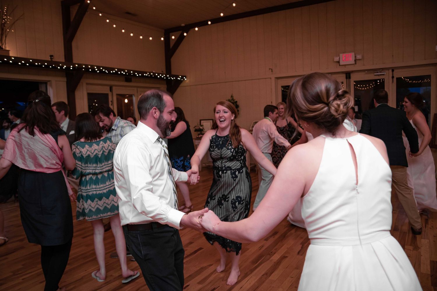 Guests dancing joyfully at a wedding reception in a warmly lit wooden venue with string lights.
