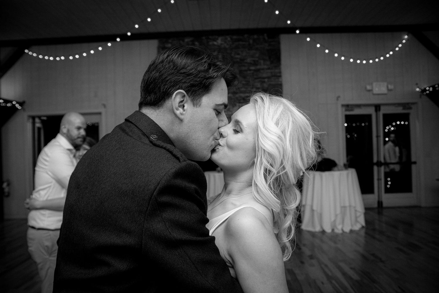 Black and white photo of a couple sharing a first dance kiss at their wedding reception with string lights in the background.