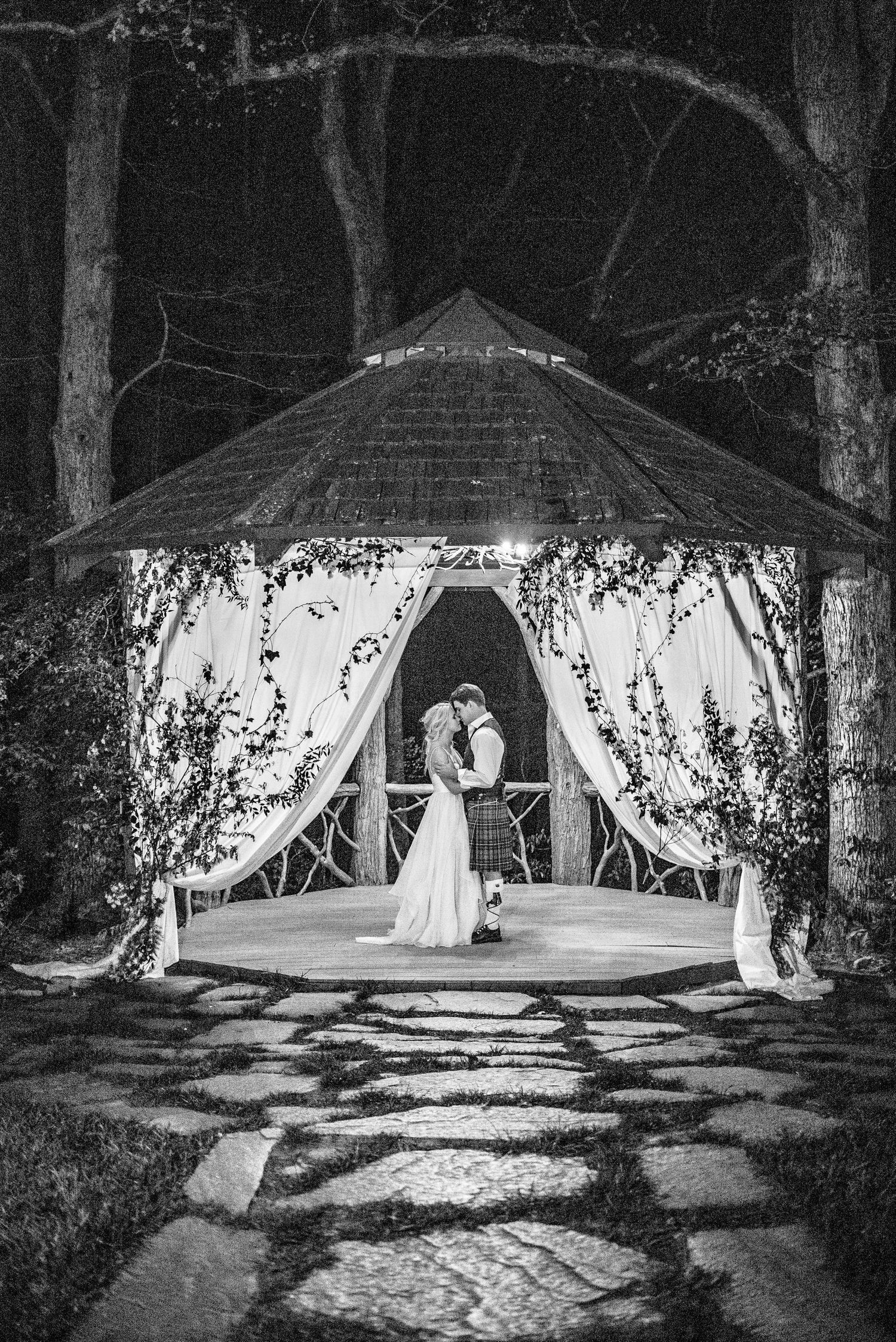 Black and white photo of a bride and groom kissing under a decorated gazebo at night.