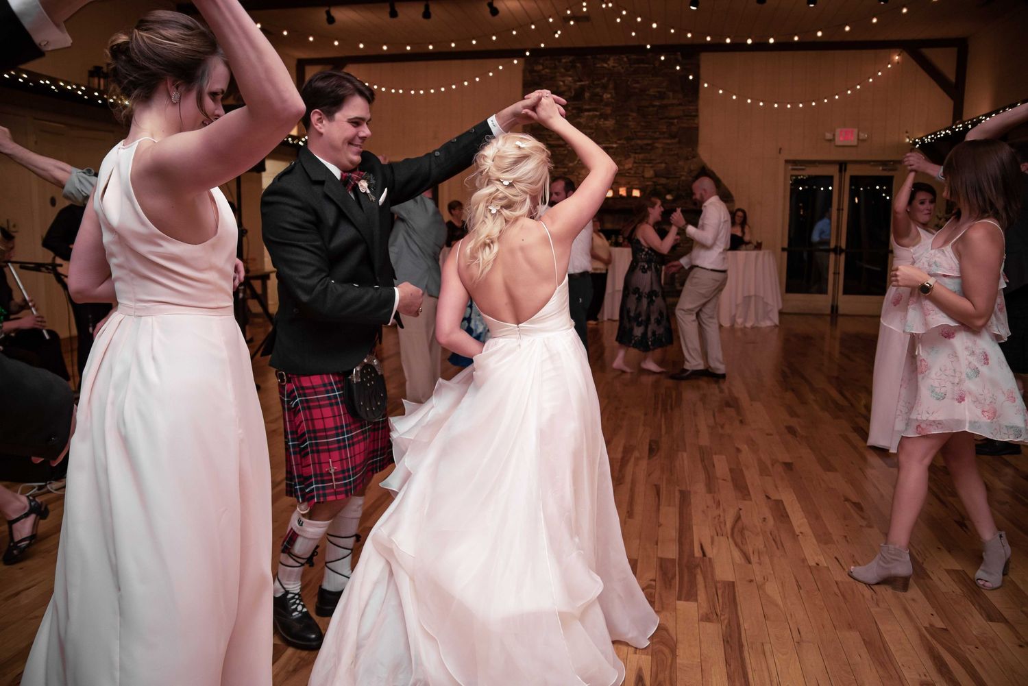 Bride and groom dancing together at wedding reception, woman in white gown, man in tartan kilt, string lights overhead.
