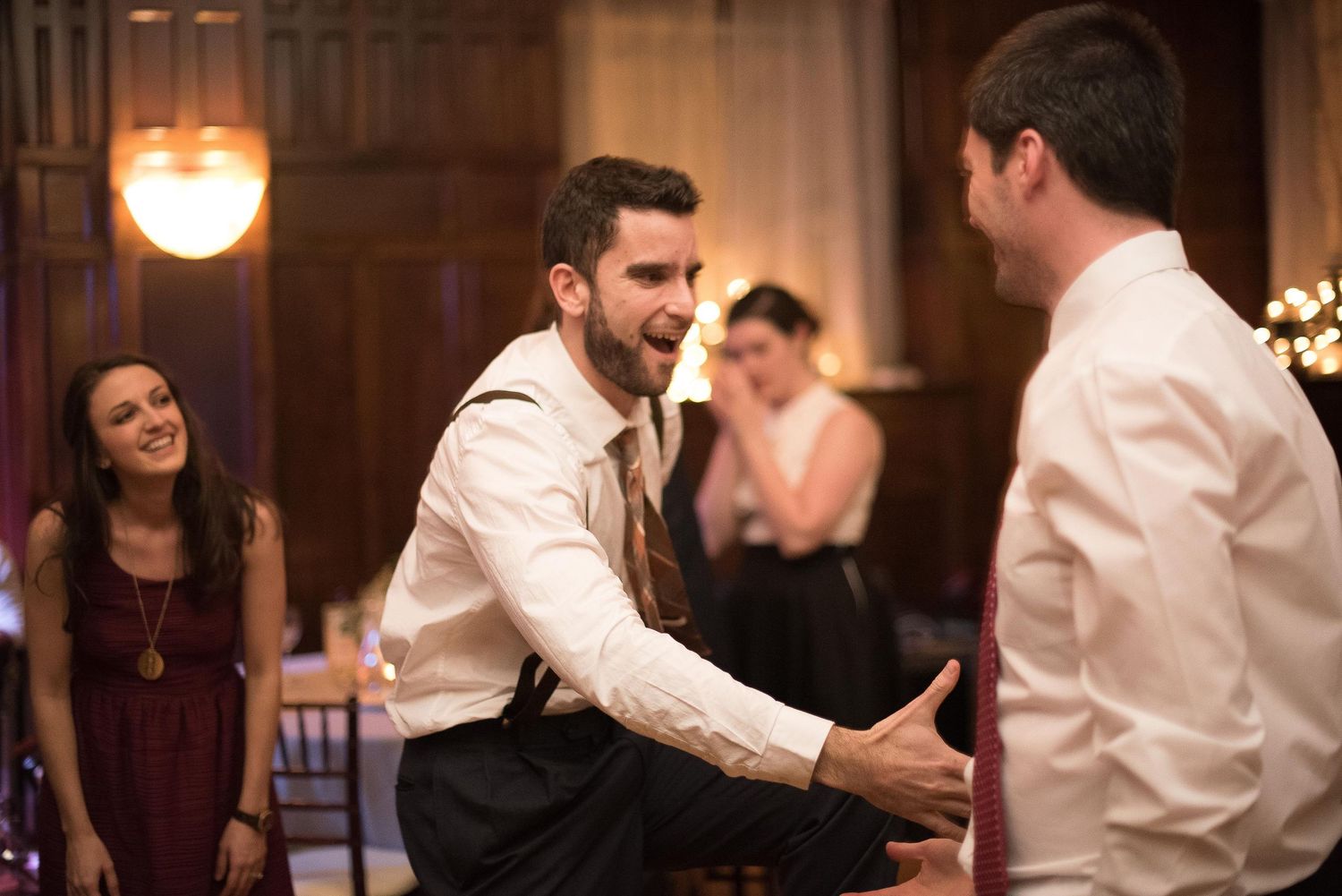Two men dancing energetically at a wedding reception while a woman in burgundy dress smiles in the background.
