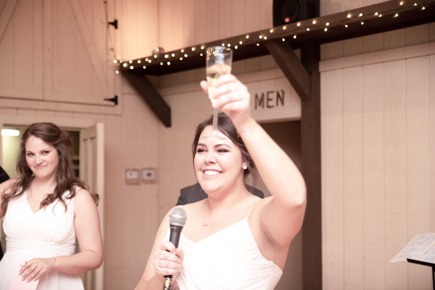 Bridesmaid raises champagne glass for toast while holding microphone at elegant wedding reception venue.