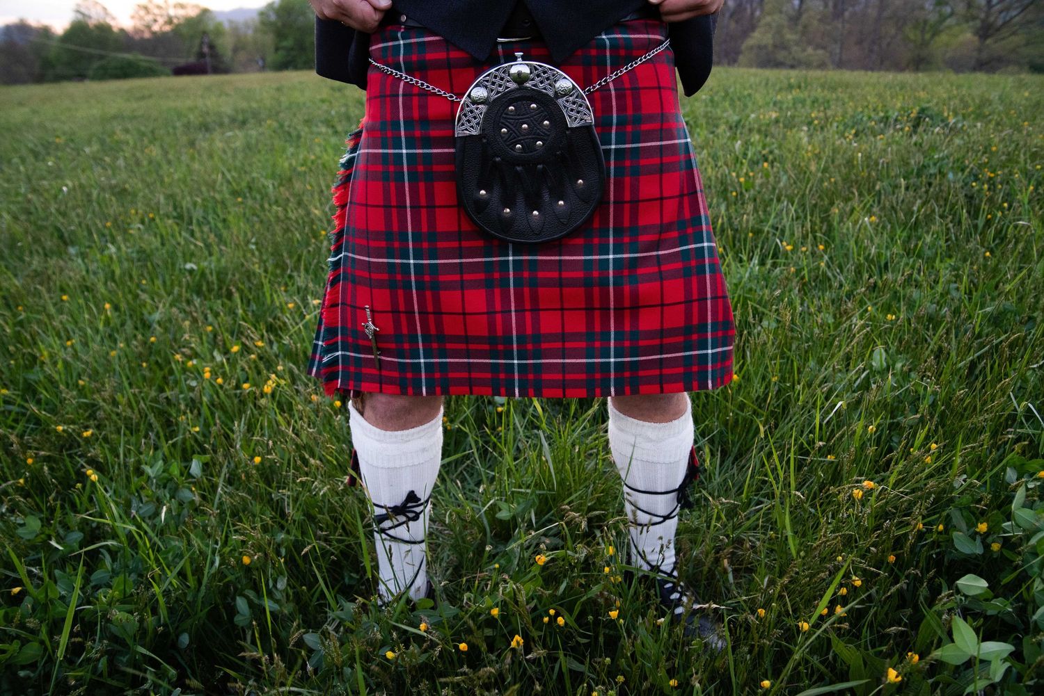 Person wearing a red tartan kilt with white socks and black shoes standing in a green meadow.