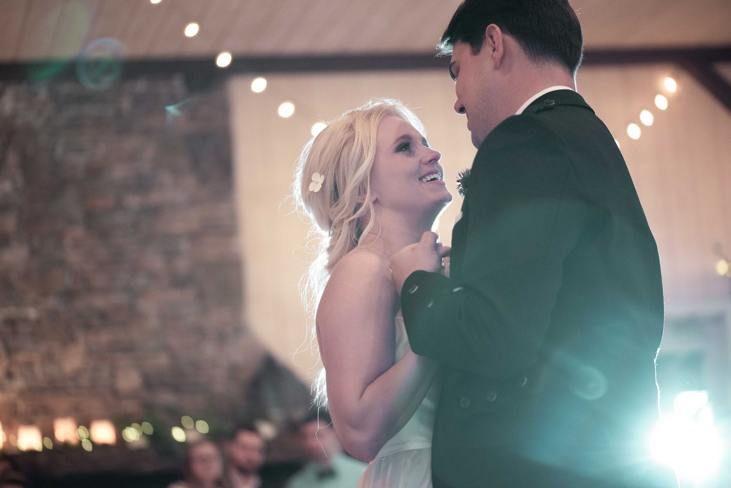 Bride and groom share their first dance in a warmly lit rustic wedding venue with string lights.