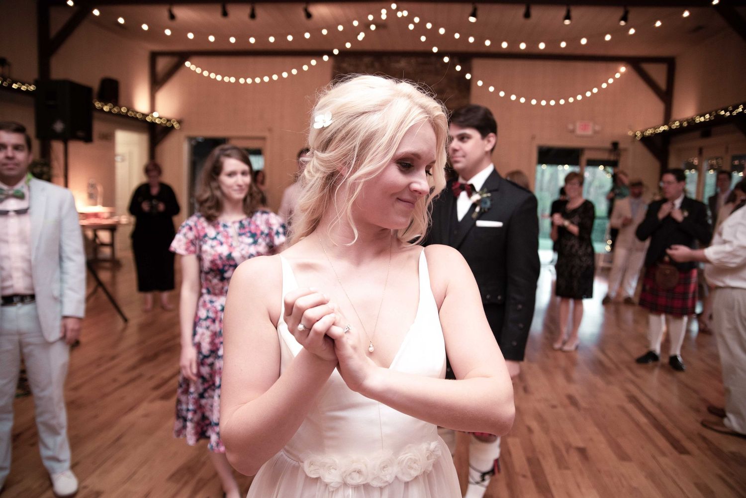 Bride in white dress clasping hands at wedding reception with guests dancing in background under string lights.