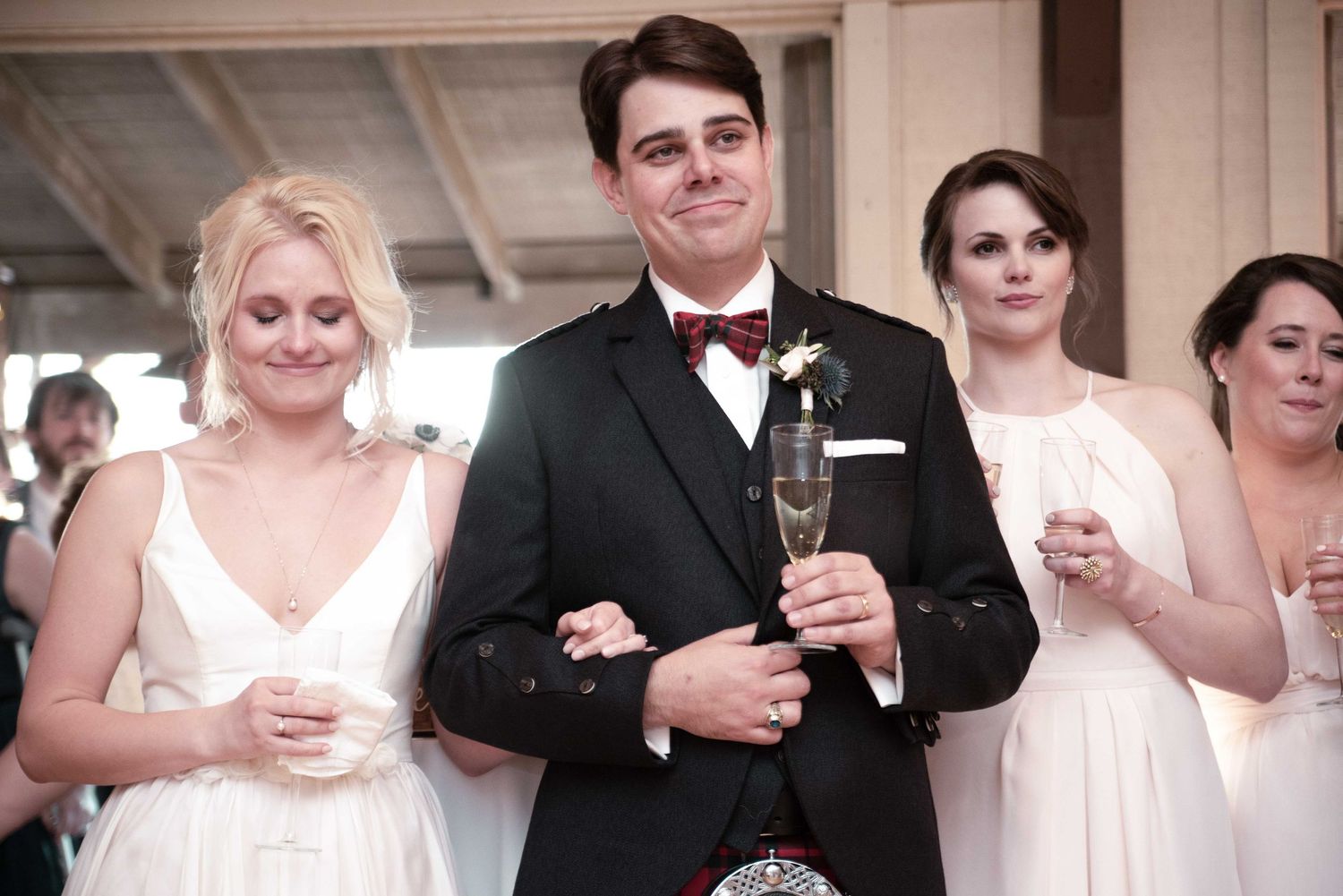 Groom in black tuxedo holding champagne flute stands between bridesmaids in white dresses at wedding reception.