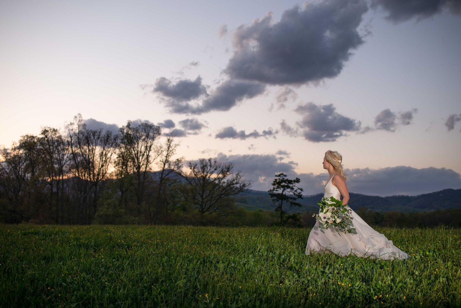 Bride in white gown holding bouquet stands in lush green field at sunset with mountains and dramatic clouds.